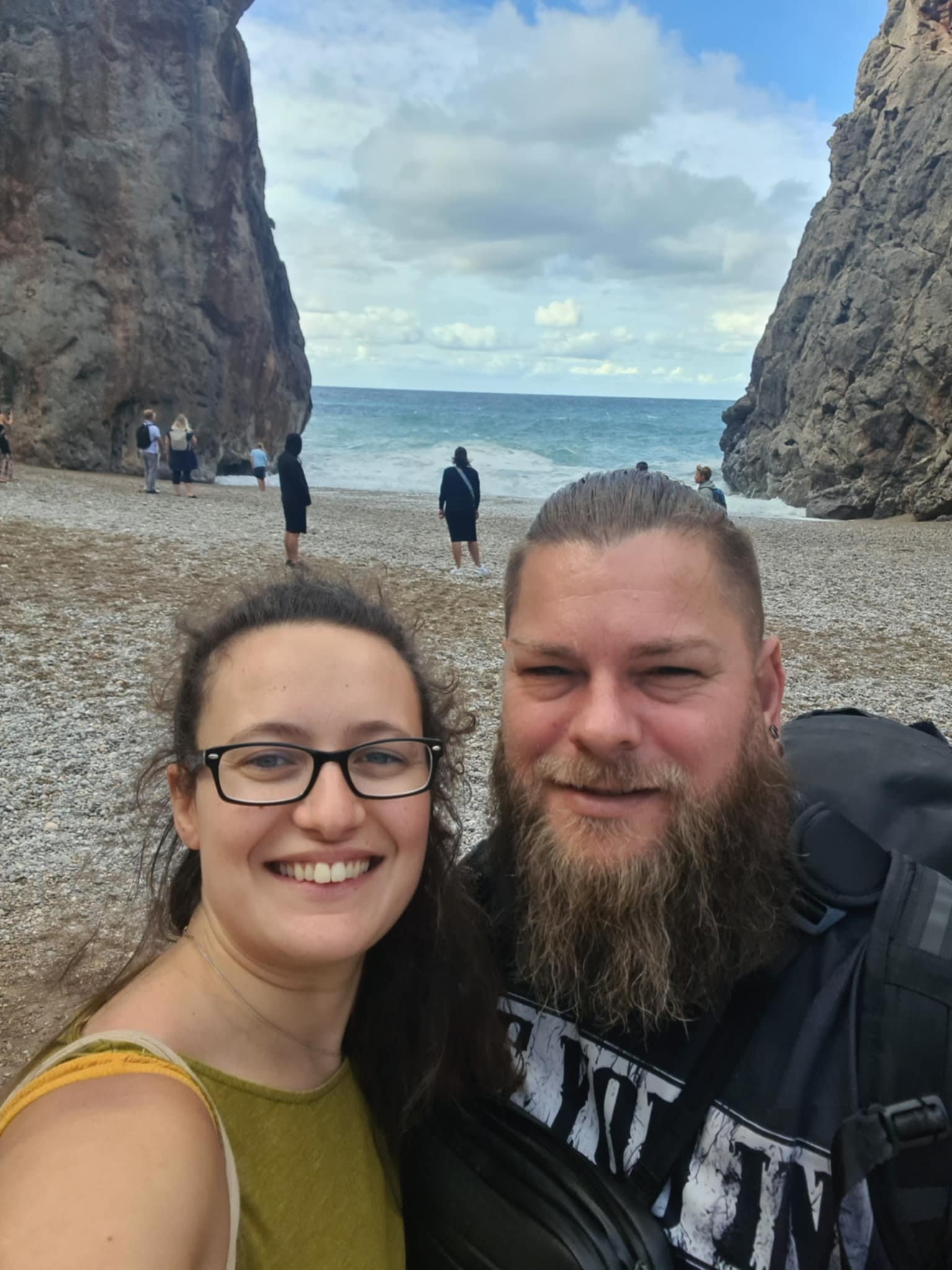 A smiling woman with glasses and a man with a beard taking a selfie on a beach with large cliffs and ocean in the background. Other people are seen walking and standing near the water.