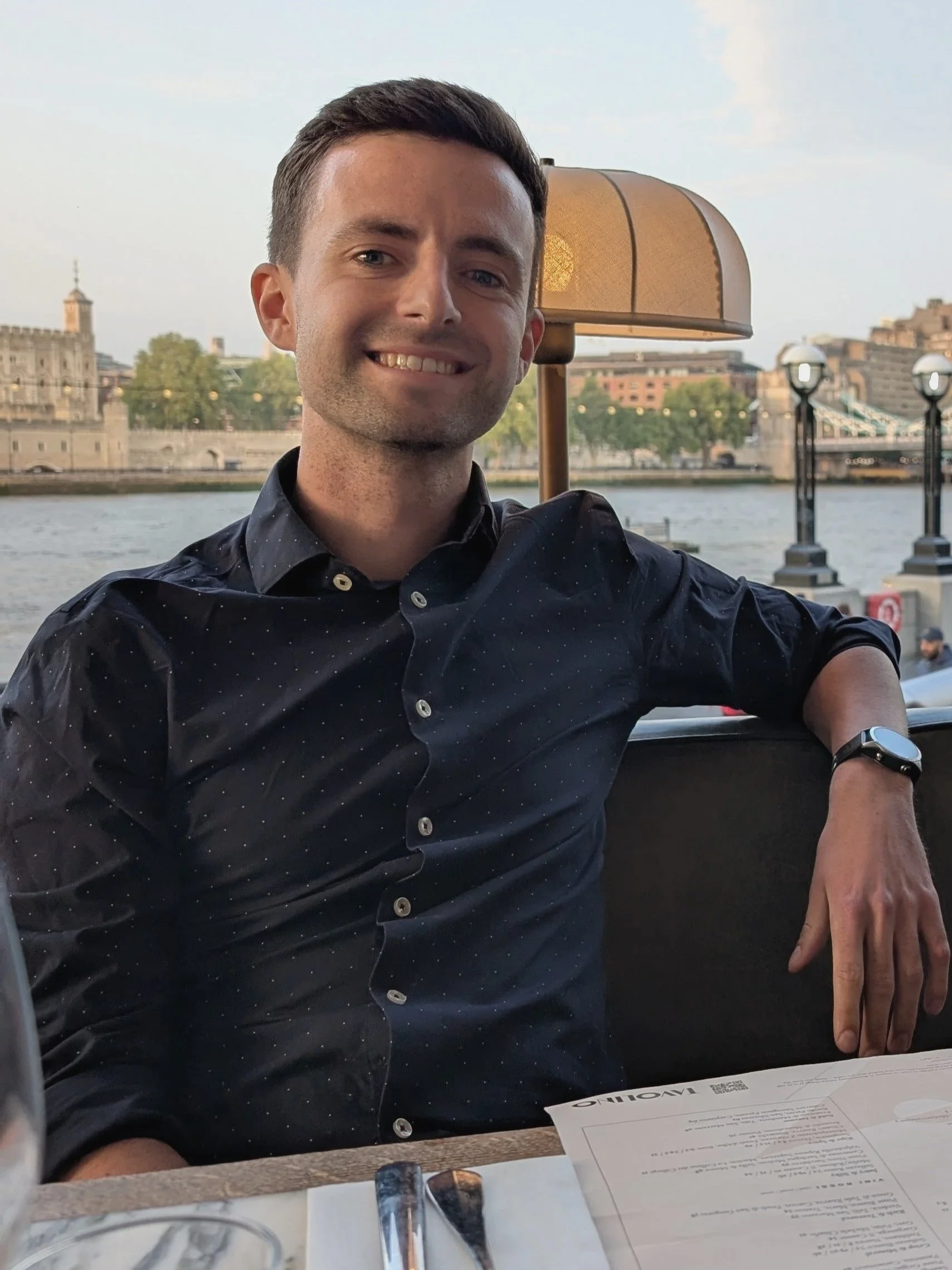 A smiling young man with short dark hair, wearing a dark button-up shirt, sitting at an outdoor restaurant table near a river with a cityscape in the background.