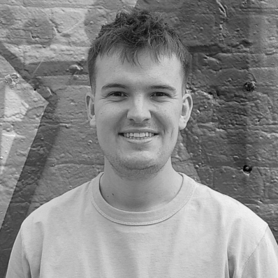 A young man with short hair and a light-colored sweatshirt smiling in front of a brick wall.