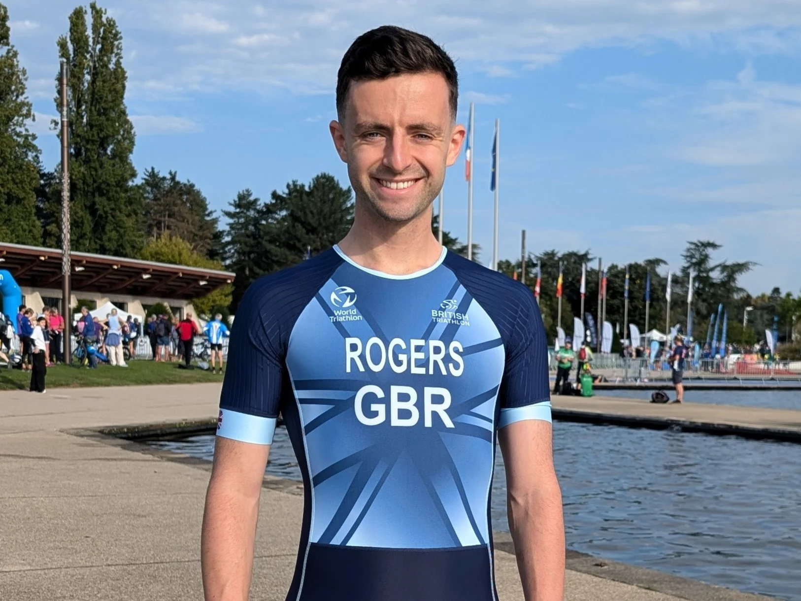 A young man in a blue triathlon uniform with "Rogers GBR" on the front, smiling outdoors near a body of water, with flags and people in the background.
