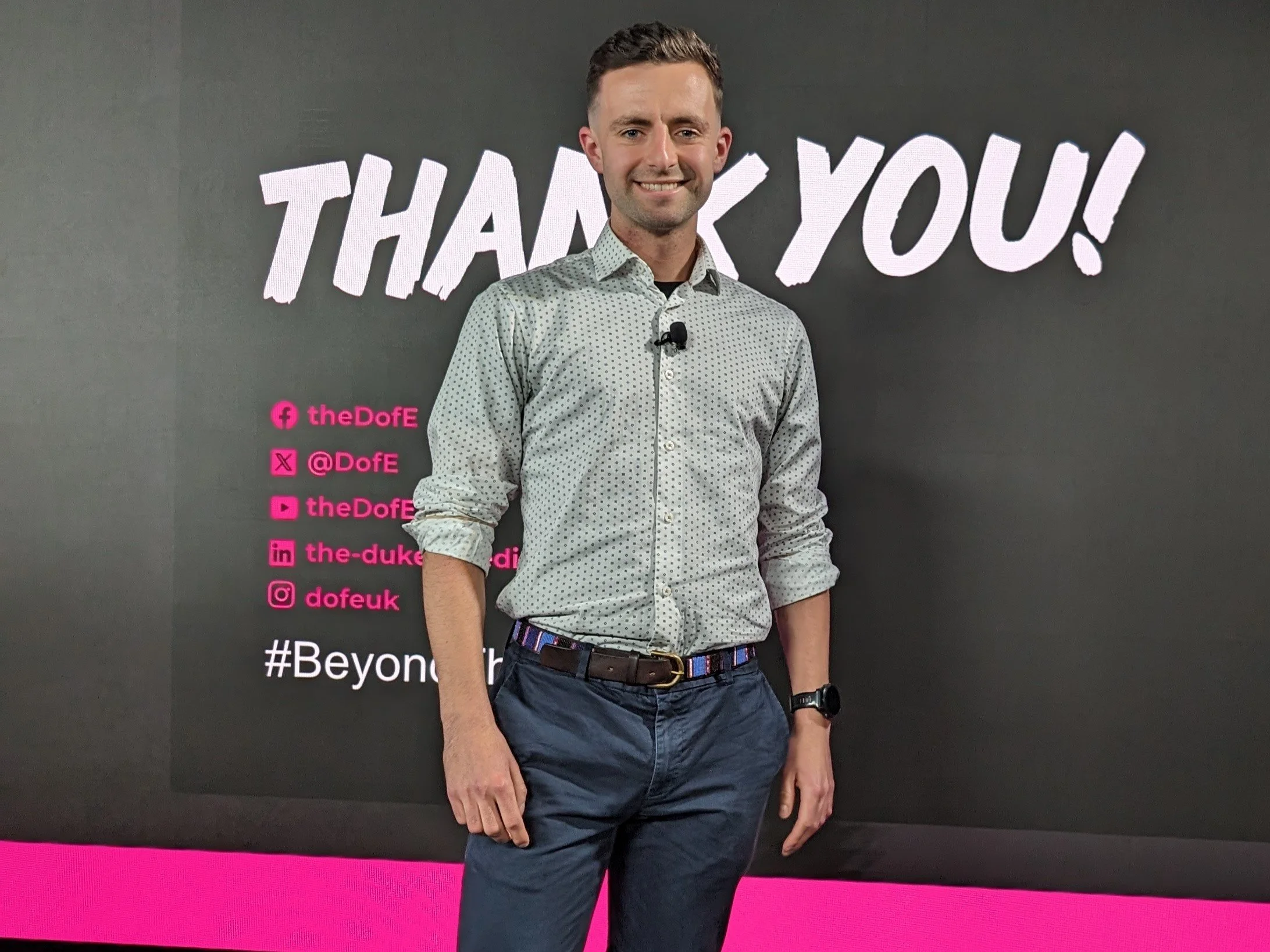 A man standing in front of a black background with pink and white text that says "THANK YOU!" and various social media handles and a hashtag.