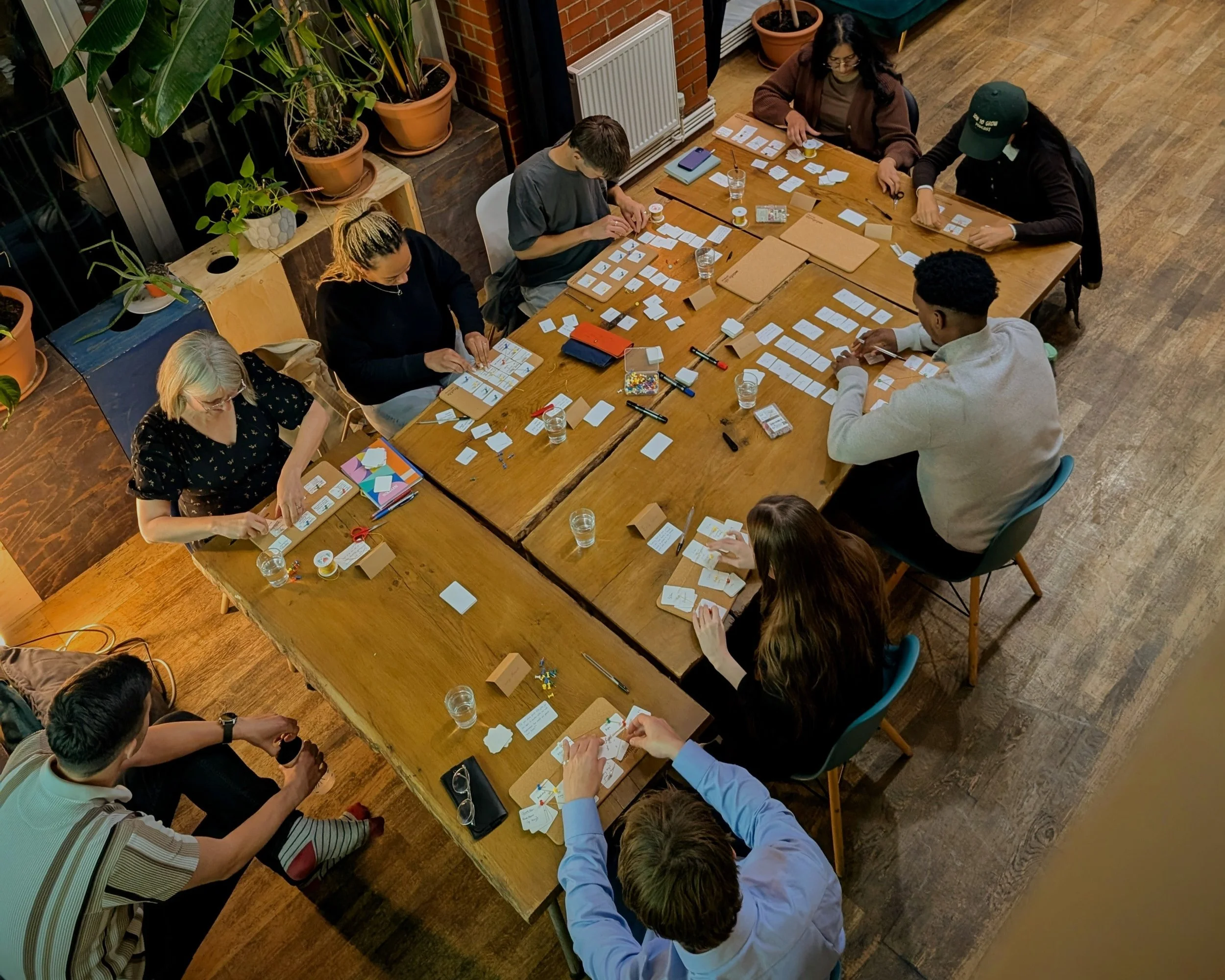 A group of diverse people sitting around large wooden tables, engaging in a card and board game activity in an indoor space decorated with potted plants, mugs, and stationery.