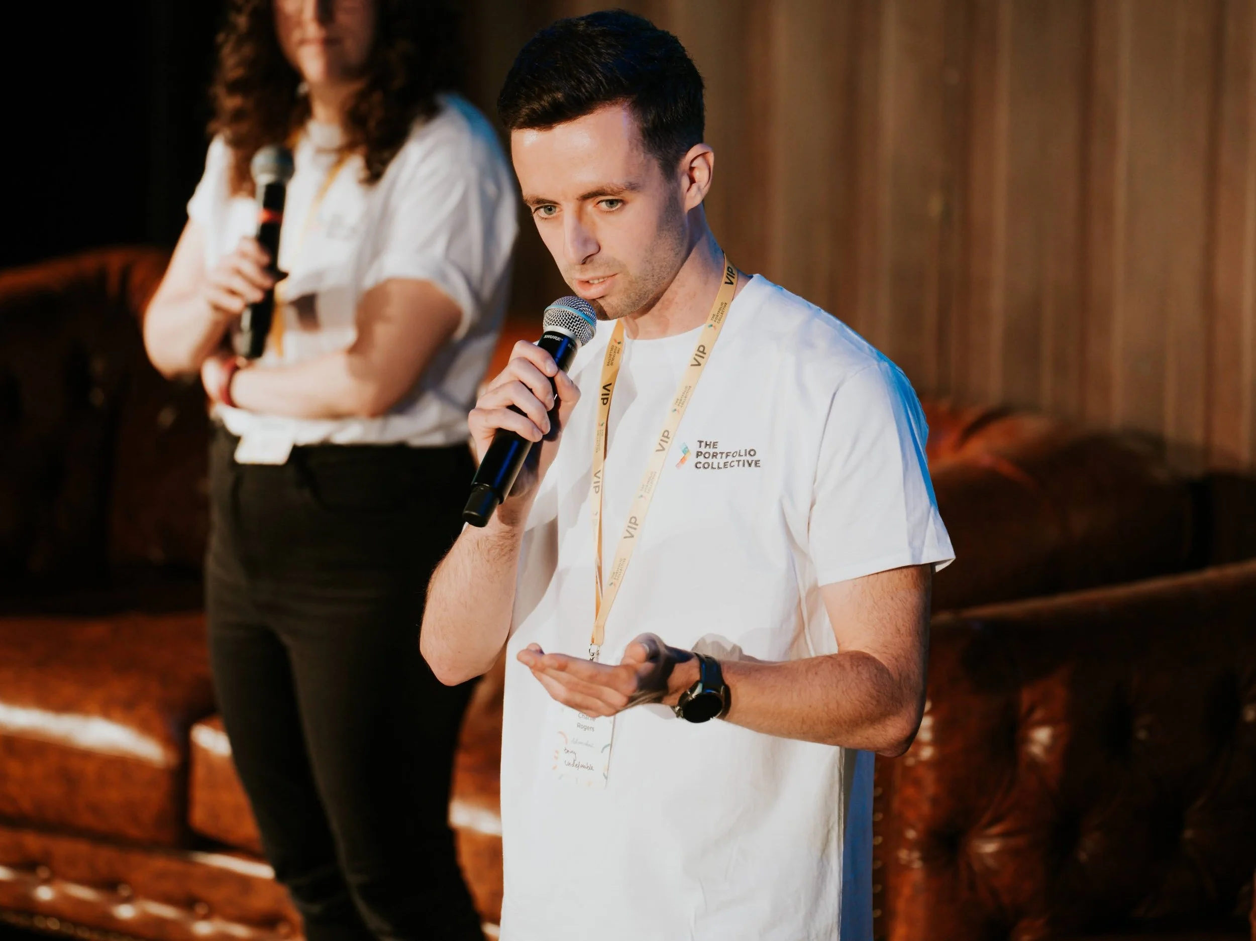 A young man wearing a white t-shirt with a logo, holding a microphone, appears to be reading or speaking, with another woman holding a microphone in the background, both on a stage with wooden paneling and a brown leather couch.