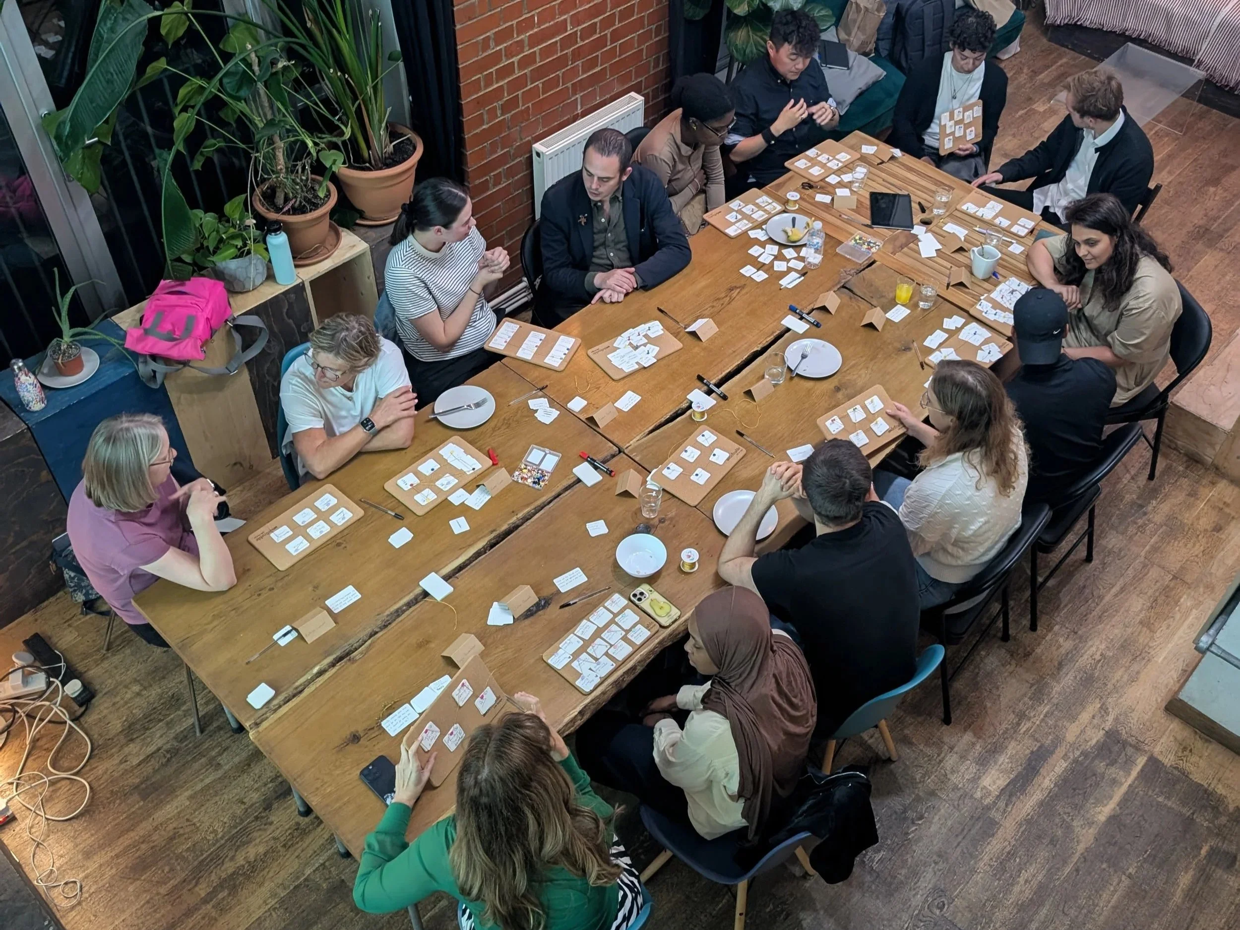 Group of diverse people gathered around a large wooden table playing a tabletop game with cards and small game pieces.