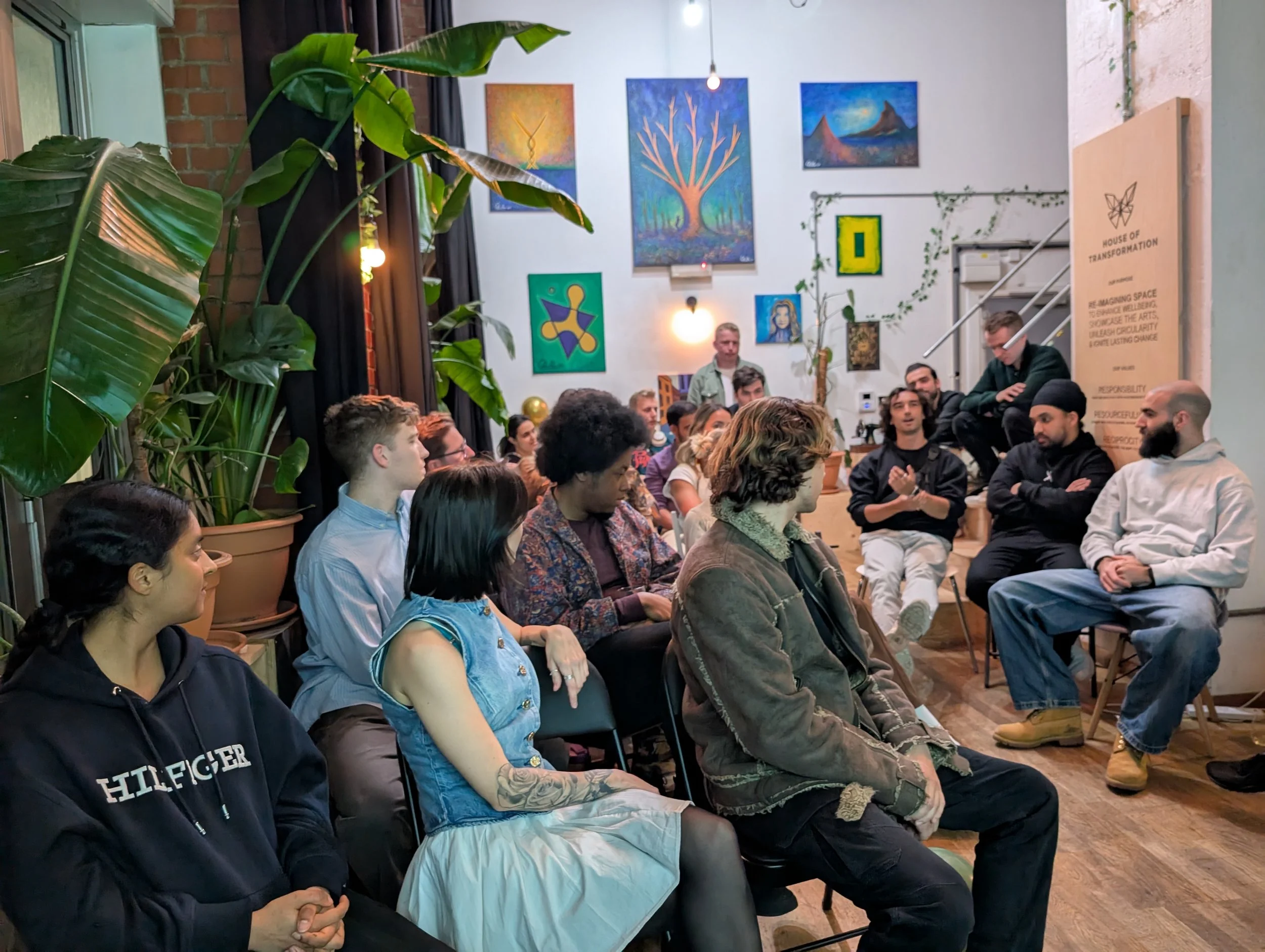 A group of diverse people seated in a room, attentively listening to a speaker at a discussion or workshop. The room has vibrant artwork on the walls and large green plants.