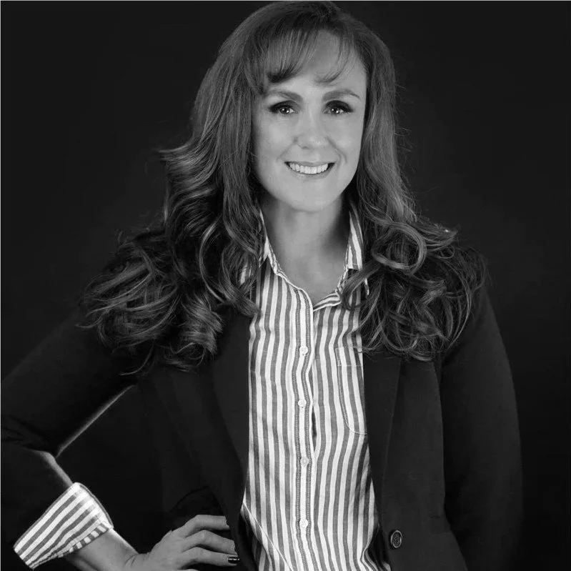 Black and white portrait of a smiling woman with wavy hair, wearing a striped shirt and dark blazer, standing against a dark background.