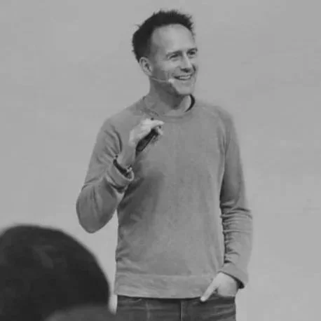 A man with dark, short hair and a smile, wearing a long-sleeve shirt and standing with his hand near his face, in front of a light-colored background.