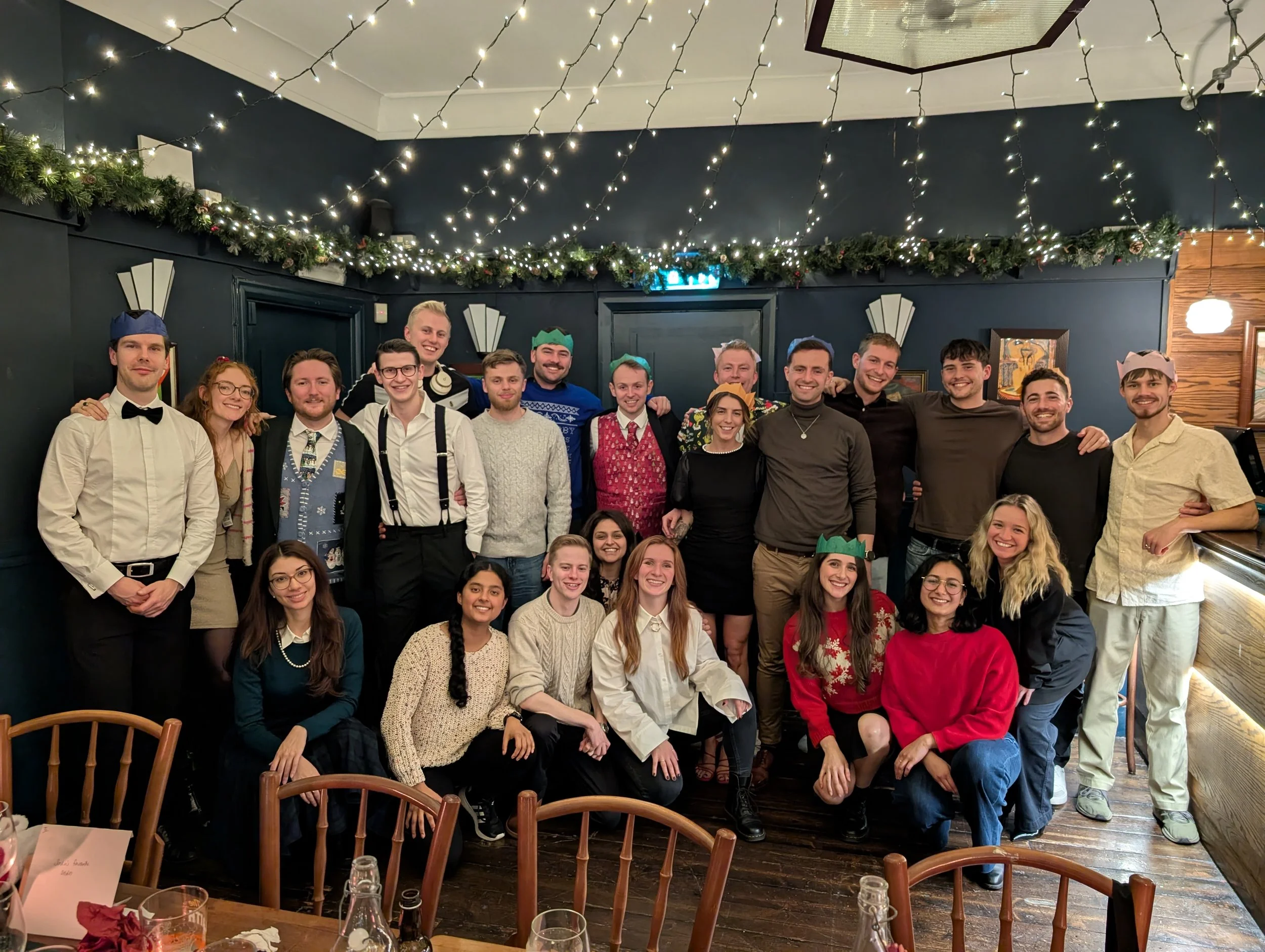 A group of 20 people gathered in a decorated indoor space for a celebration, likely Christmas, with string lights and greenery along the ceiling, and some wearing paper crowns and festive attire.