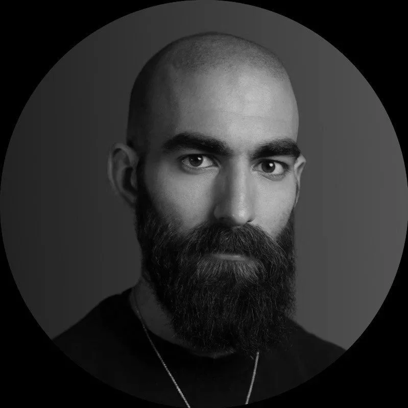 Black and white portrait of a man with a shaved head, thick beard, intense eyes, and wearing a necklace, against a dark background.
