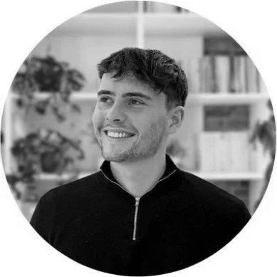 A young man with short, dark hair and a smile, wearing a zip-up black jacket, in an indoor setting with bookshelves and plants in the background.