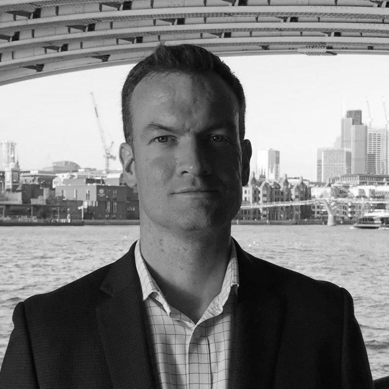 Black-and-white photo of a man in a suit standing outdoors near a river with a cityscape in the background, featuring tall buildings and construction cranes under a bridge.
