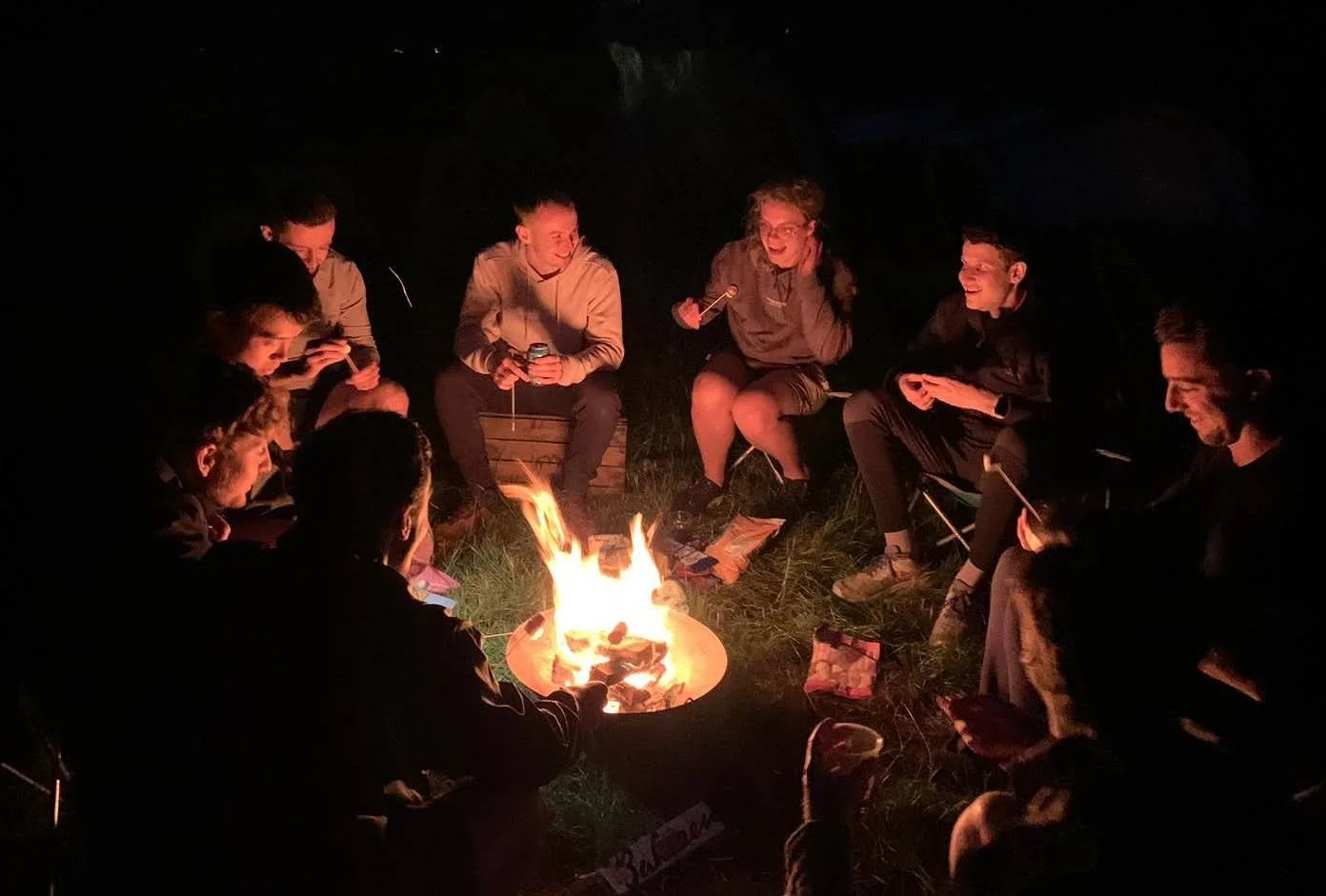 Group of people sitting around a campfire at night, some holding drinks and talking, with dark surroundings.