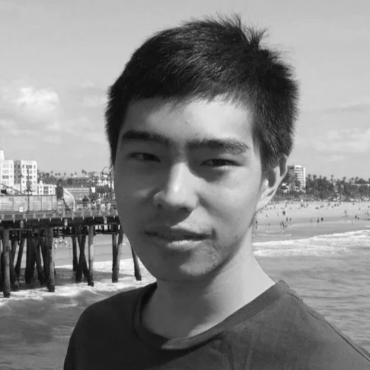 A young Asian man with short black hair smiling at the camera on a beach with a pier and buildings in the background.