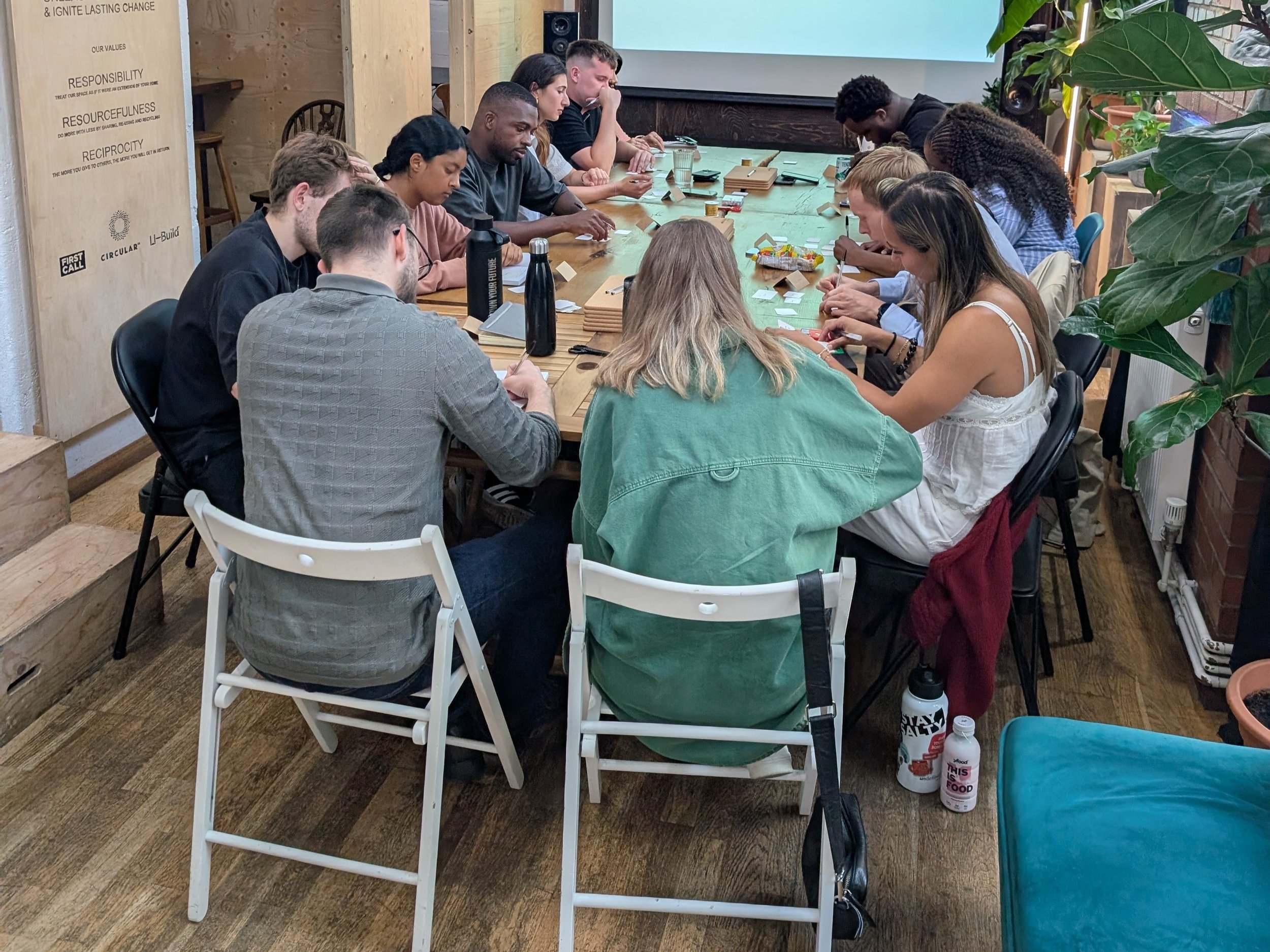 A group of people participating in a board game or card game session around a rectangular wooden table in a cozy, eco-friendly indoor setting with plants and wooden accents.