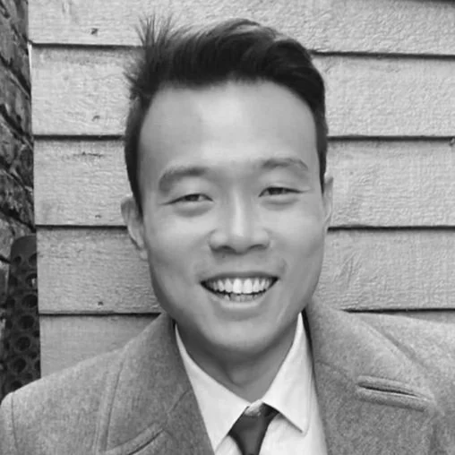 A smiling man in a suit and tie posing outdoors against a wooden wall.