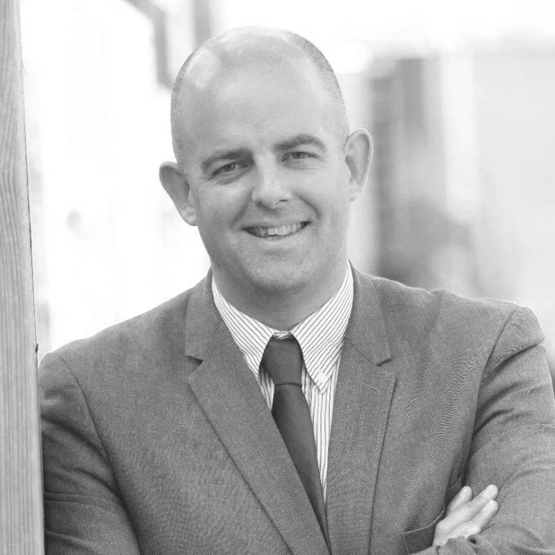 Black and white portrait of a smiling man wearing a suit and tie, standing outdoors.