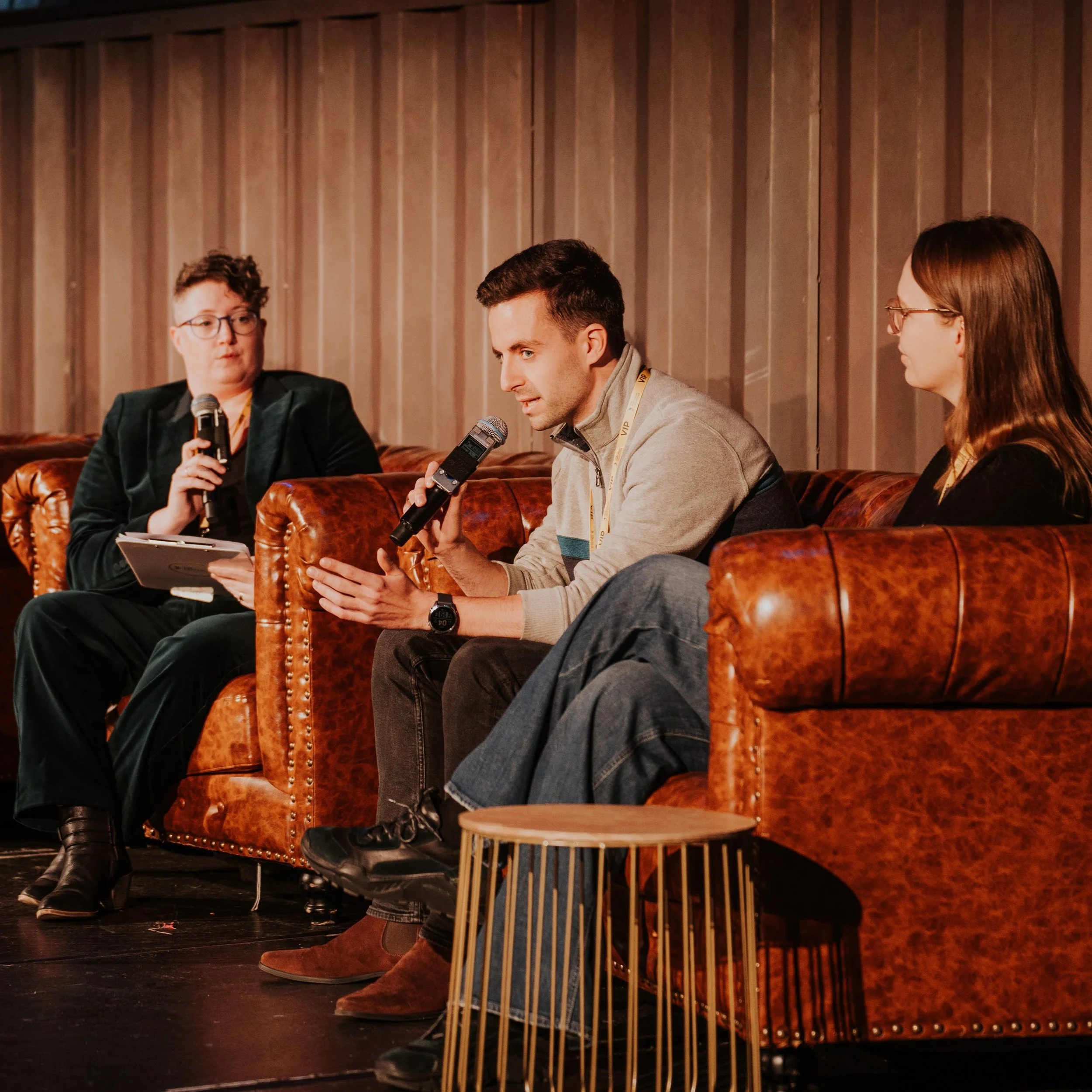 A panel discussion with three people seated on brown leather couches, one holding a microphone and speaking, amid a warm, wood-paneled background.