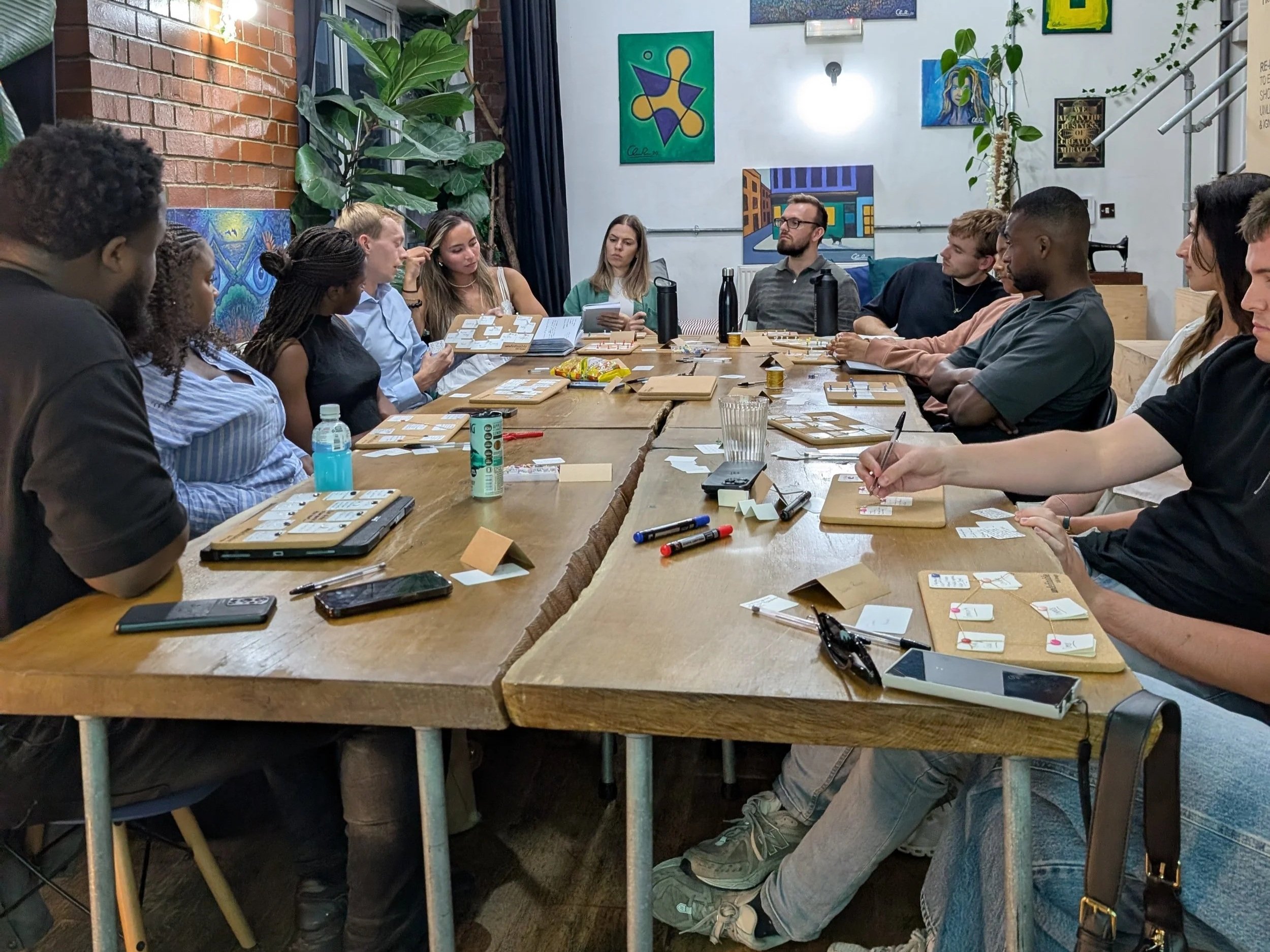 A diverse group of people sitting around a large wooden table in a casual meeting or game night setting, engaging with various cards, game pieces, and notebooks, with artwork on the walls.