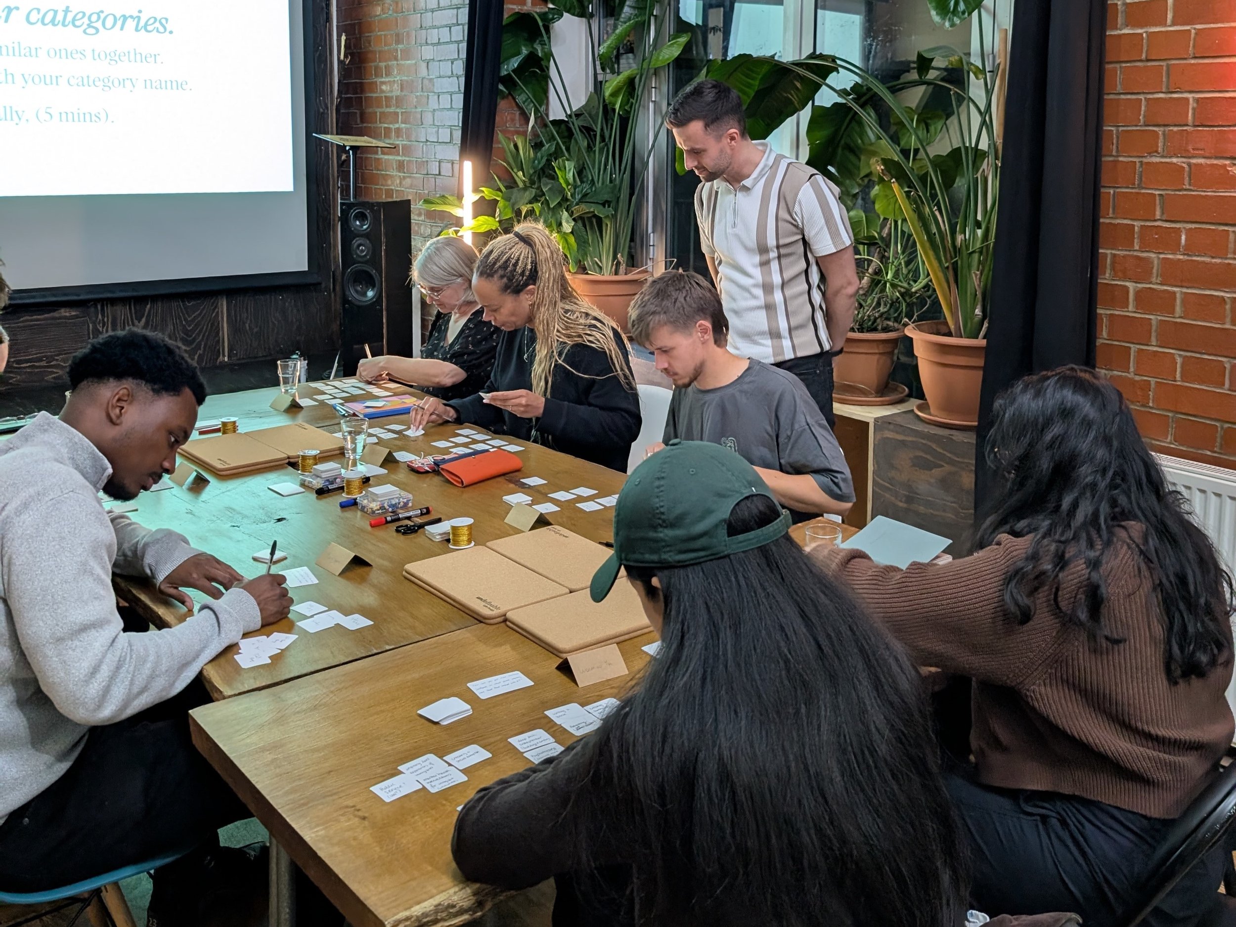 A group of people sitting at a long wooden table working on a collaborative project with papers, cards, and writing tools. There are large potted plants, a brick wall, and a projector screen in the background.