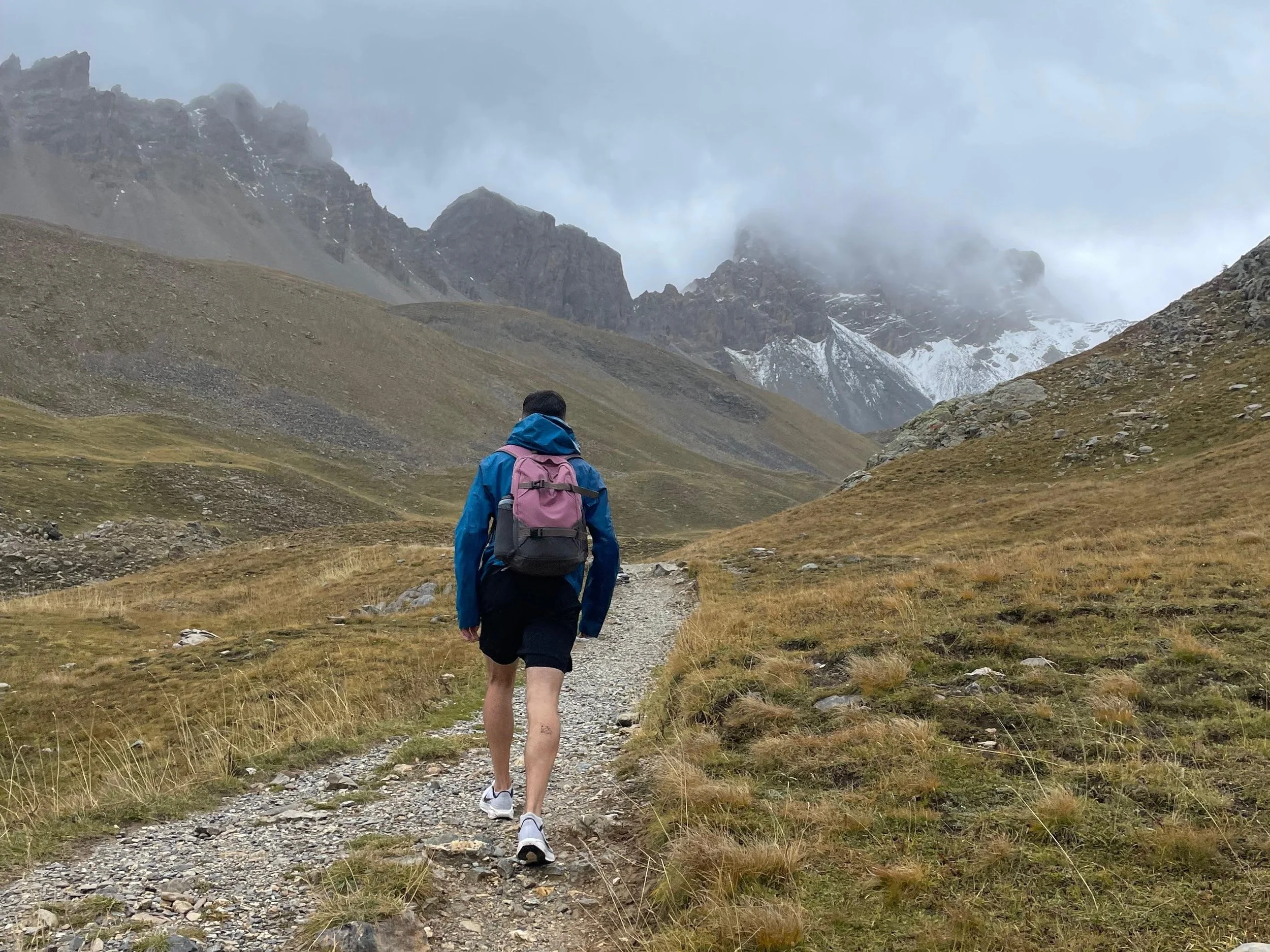 A person hiking on a rocky trail in a mountainous landscape with grassy slopes and snow-capped peaks in the background.