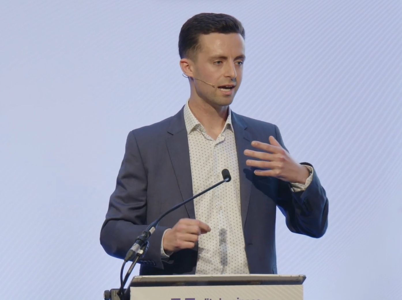 A man speaking at a podium with a microphone, wearing a dark blue suit, white shirt, and a headset microphone, gesturing with his right hand.