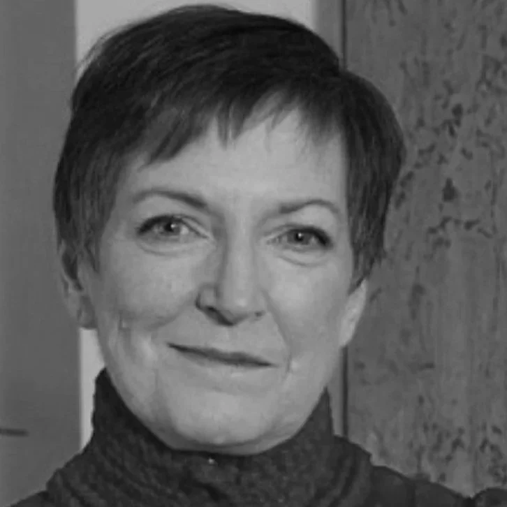A black and white close-up photograph of a smiling woman with short dark hair, wearing a dark turtleneck sweater, against a background of a wooden surface.