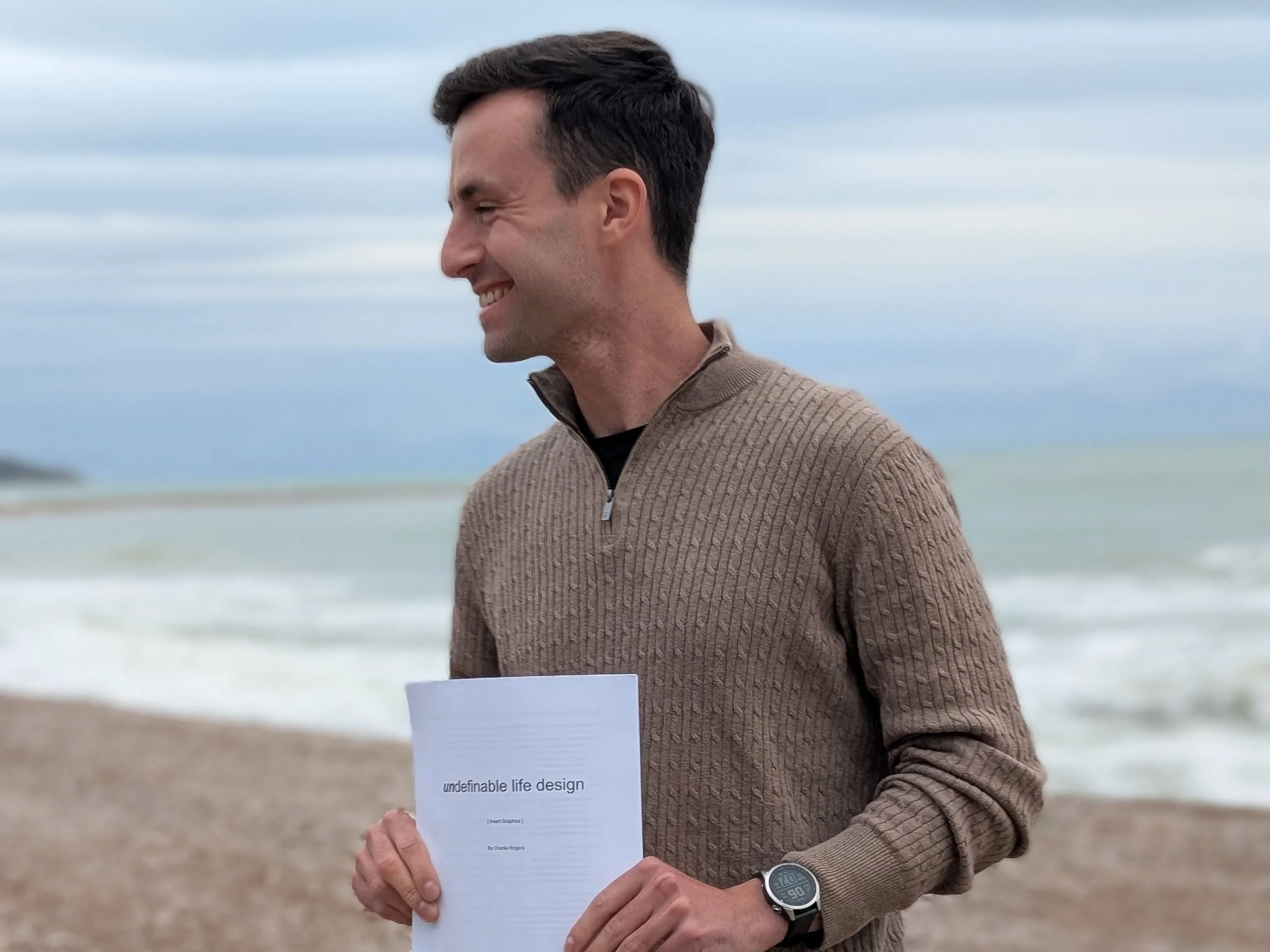A man with dark hair smiling, wearing a military brown sweater, holding a printed document titled 'undefinable life design' while standing on a beach with waves and cloudy sky in the background.
