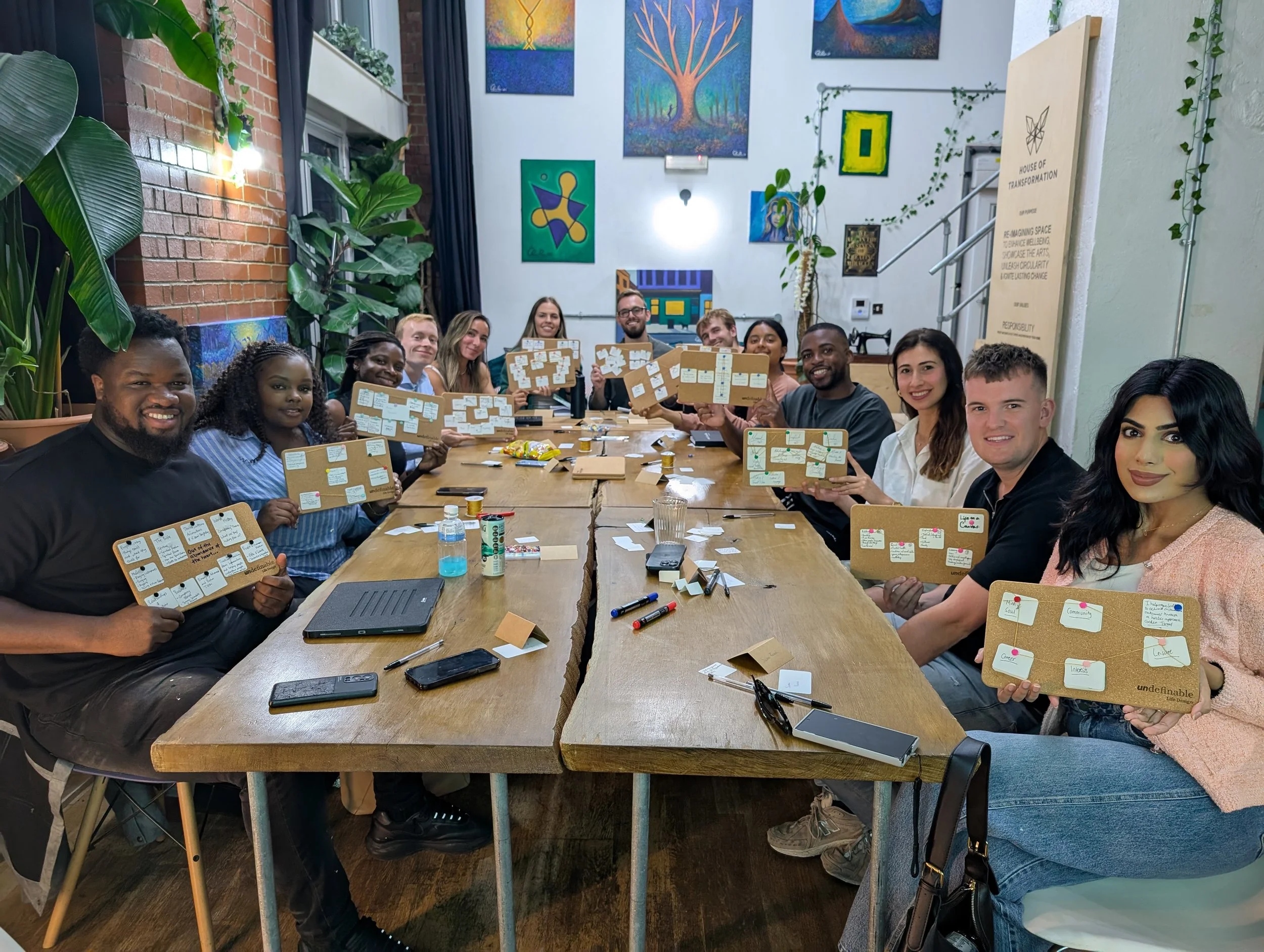 Group of diverse people sitting around a long wooden table, holding up bulletin boards with notes, in a room with colorful artwork and plants.