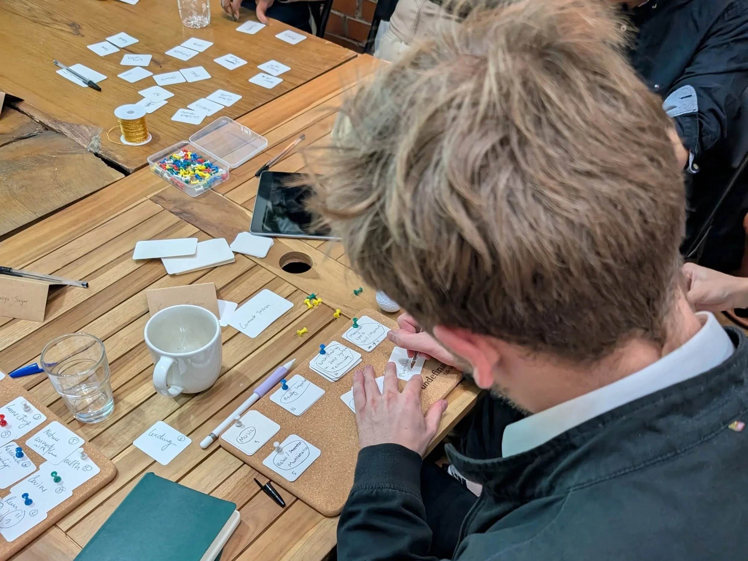 A man with brown hair and a black jacket is working at a wooden table, arranging white cards with handwritten notes attached with colorful push pins. The table has various items including a cup, a glass of water, pins, a white pen, a notebook, and a container of small colored push pins. In the background, there are more cards, markers, tools, and possibly people engaged in a similar activity.