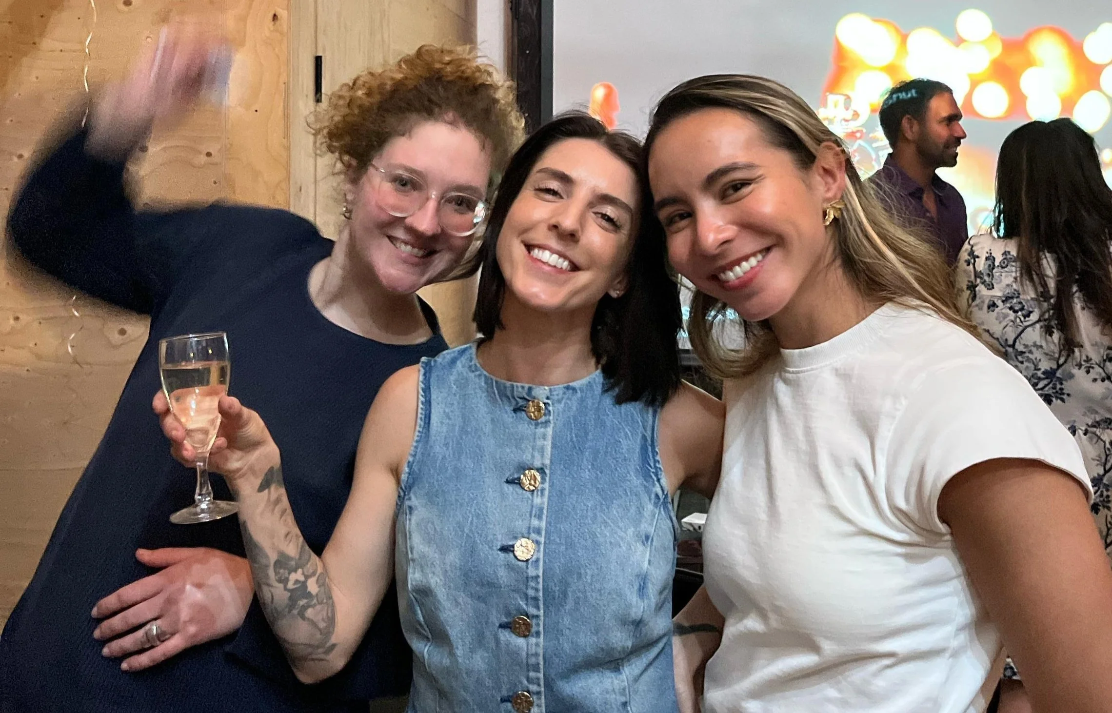 Three women smiling and posing together at a social gathering with a wooden wall background and other people in the distance. One woman holds a glass of champagne.