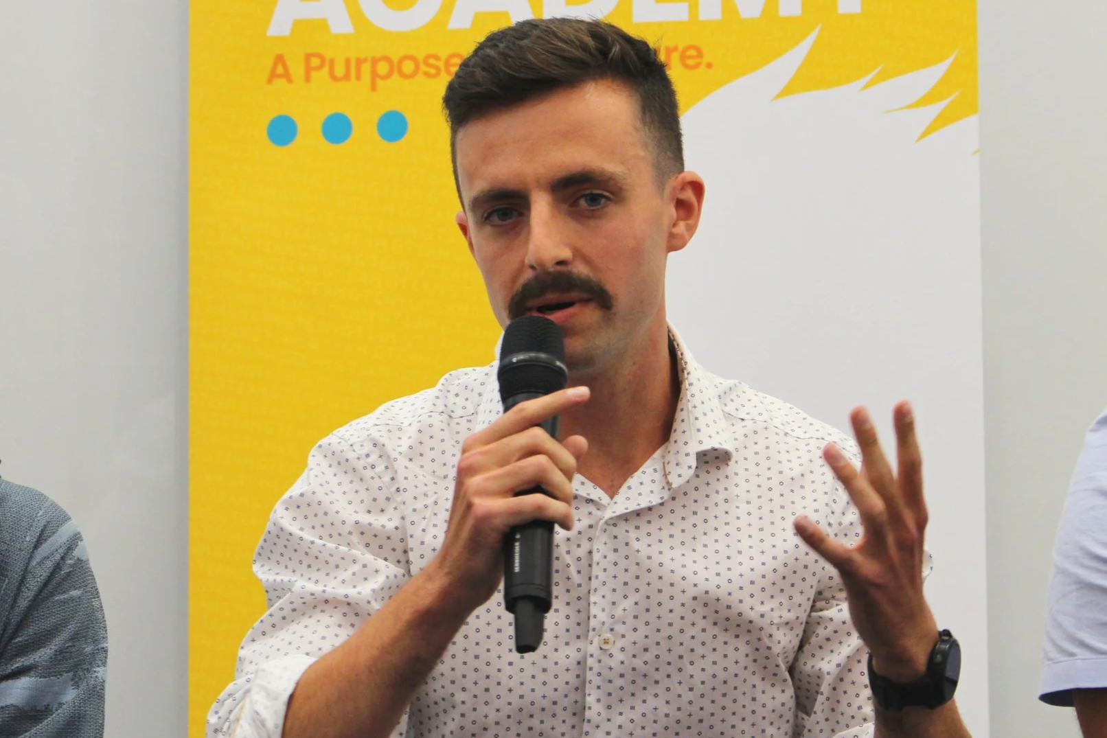 A man with dark hair, a mustache, and a goatee, wearing a white shirt with small black and purple dots, is speaking into a black microphone. He is gesturing with his left hand, and is wearing a watch on his left wrist. Behind him is a yellow background with text and blue dots, part of a larger banner or poster.