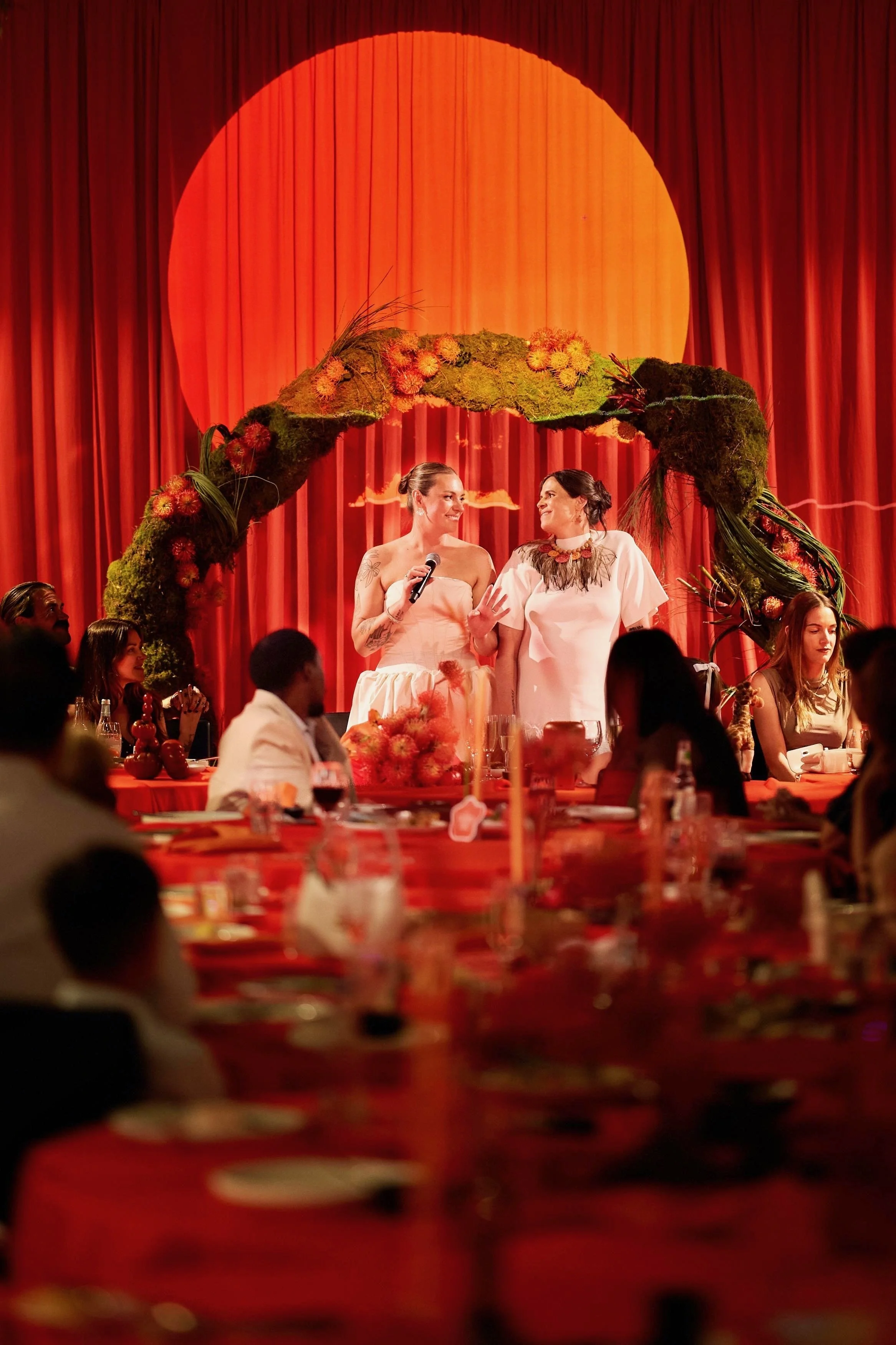 Two women stand on a stage under a large orange circle with a red curtain backdrop, speaking to an audience at a formal event with red tablecloths and table settings.