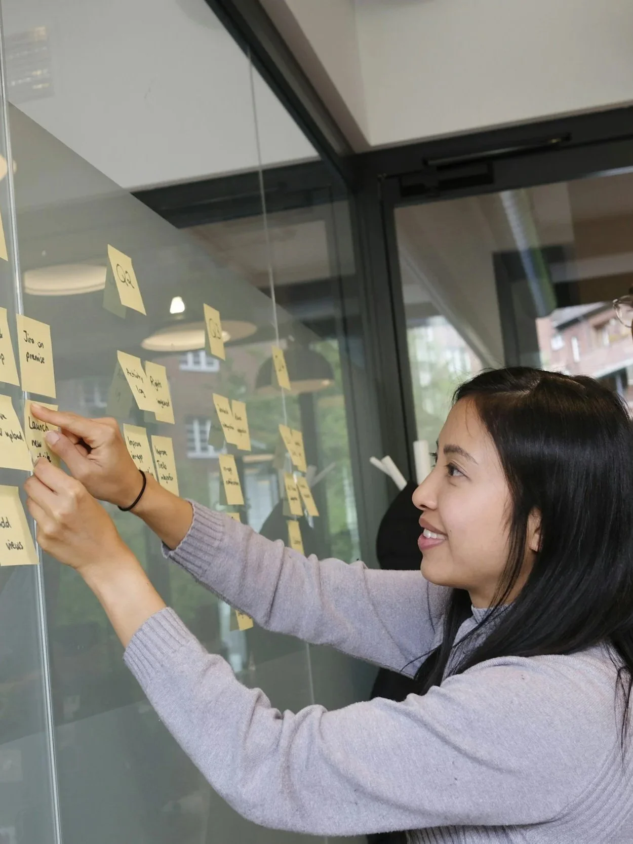 Una mujer coloca notas adhesivas en una pared de vidrio en una oficina, mientras que en el fondo hay ventanas y un edificio visible desde el interior.