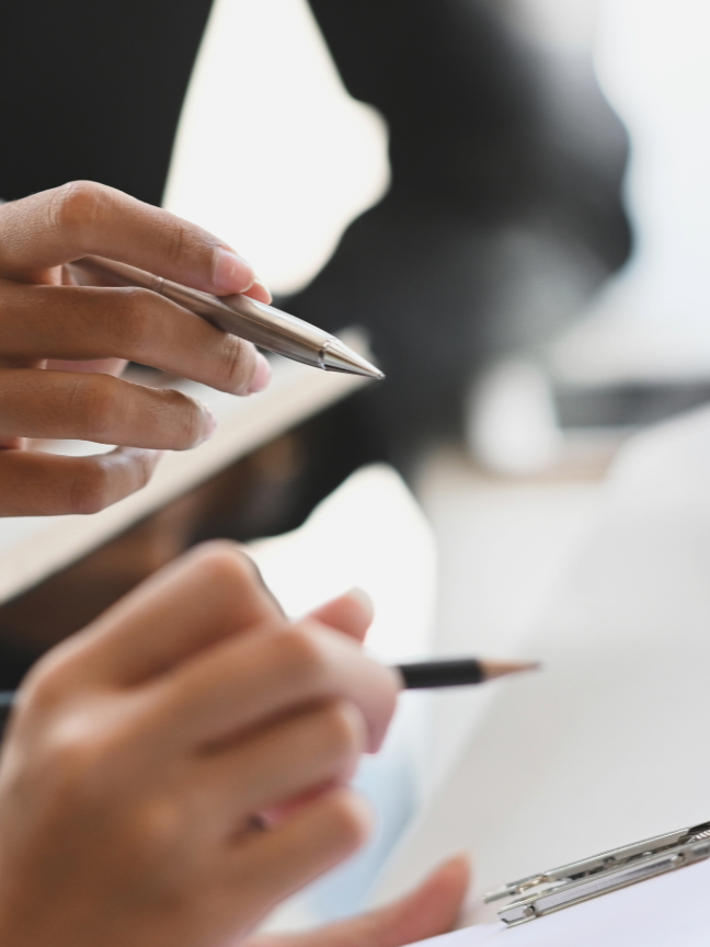 Dos personas tomando notas en una reunión o en un entorno de trabajo, usando bolígrafos y papeles.