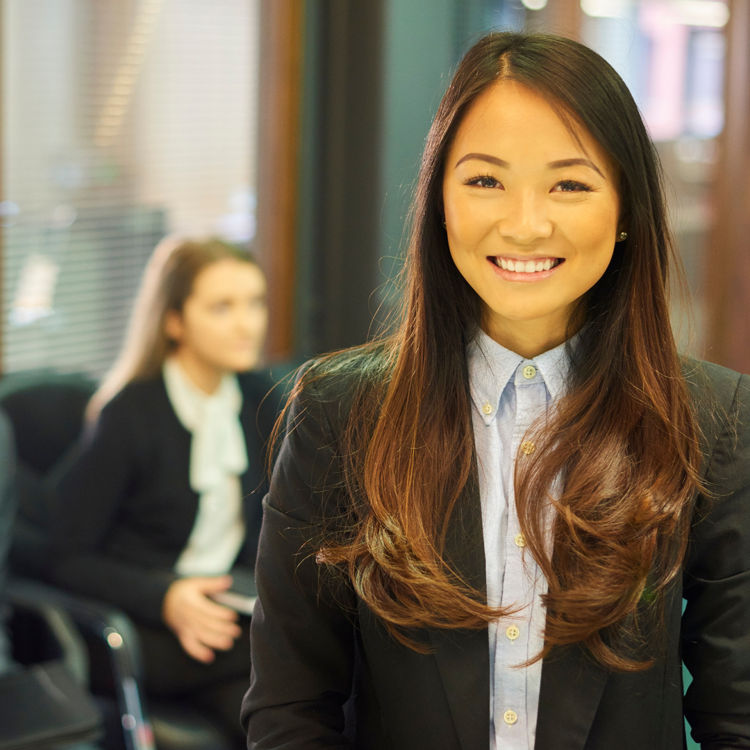 A smiling Asian woman in business attire in an office environment, with another woman blurred in the background.