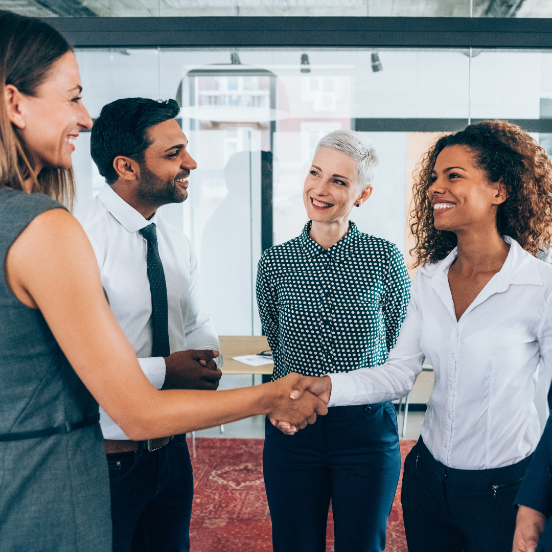 A group of four diverse professionals at a business meeting, with two women shaking hands and smiling.