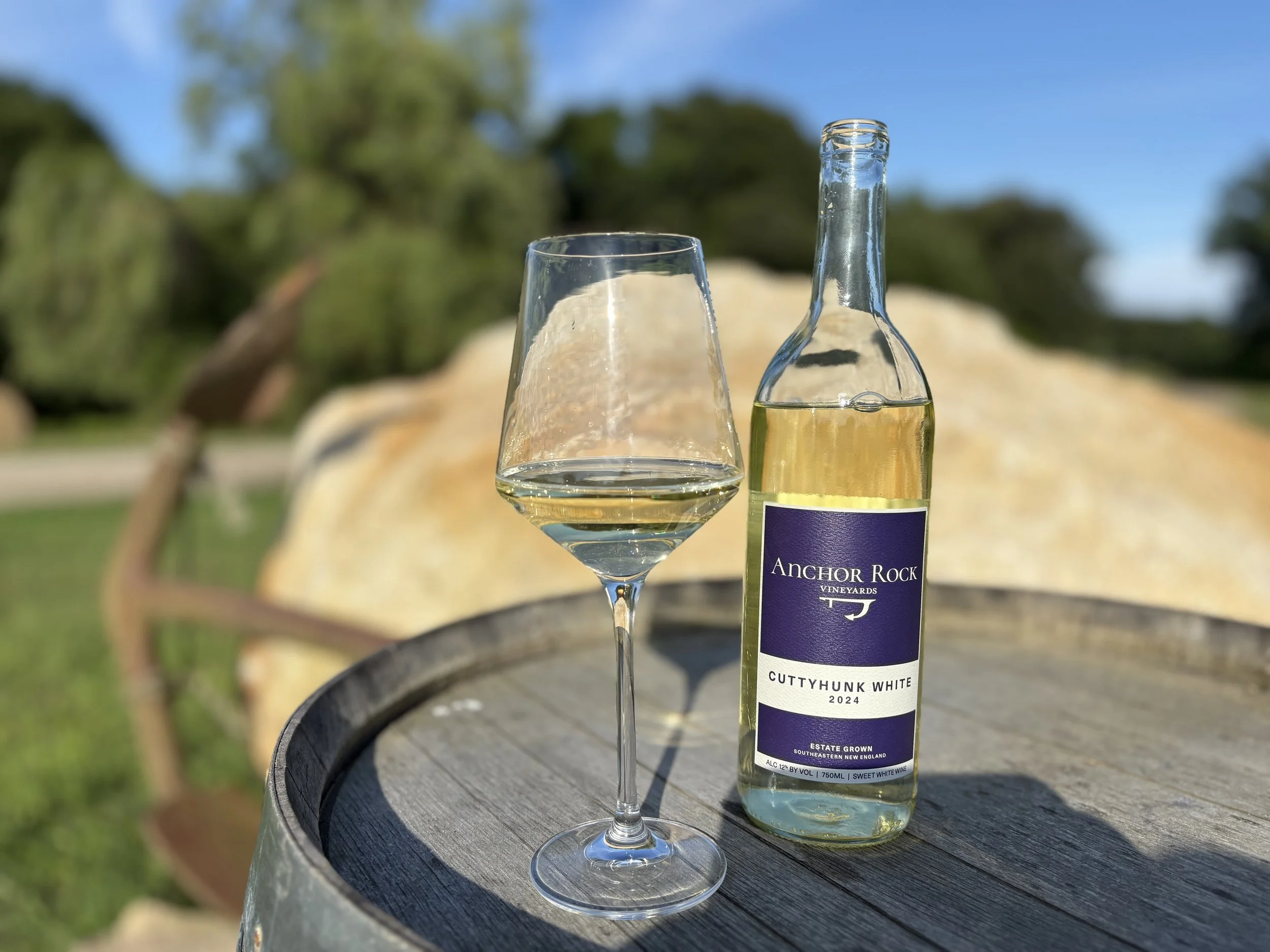 A wine glass filled with white wine and a bottle of Anchor Rock vineyards Cutthunk White 2024 estate grown wine on a round wooden surface outdoors with blurred green trees and a blue sky in the background.
