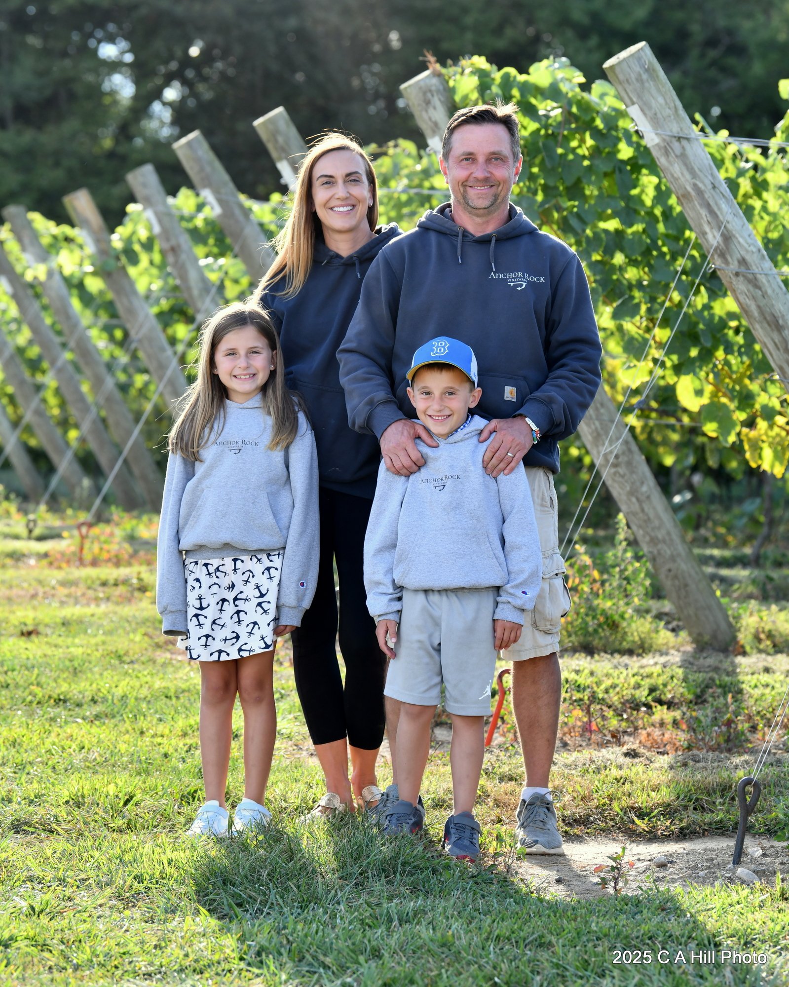 A happy family of five standing outdoors in a vineyard with green vines and wooden posts. The family includes a woman, two girls, and two boys. They are smiling and wearing casual clothes, with some in matching sweatshirts that say 'Anchor Rock.' The sunlight filters through the trees in the background.