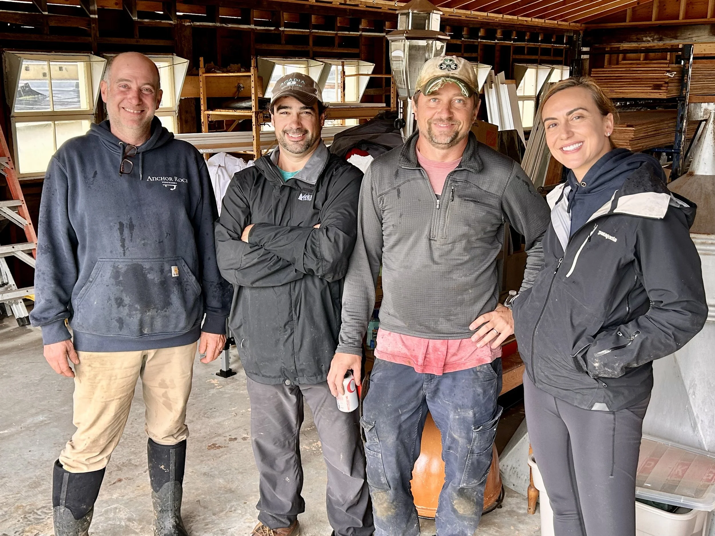Four people standing inside a wooden workshop, smiling at the camera. They are dressed casually, some in jackets and rubber boots, with shelves and various wooden materials in the background.