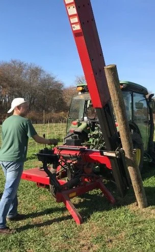 A person wearing a white cap and green shirt using a tree spade to transplant a tree outdoors. The tree spade is attached to a red and black tractor, with a large wooden post leaning against it. The background shows a grassy field and some trees under a clear blue sky.