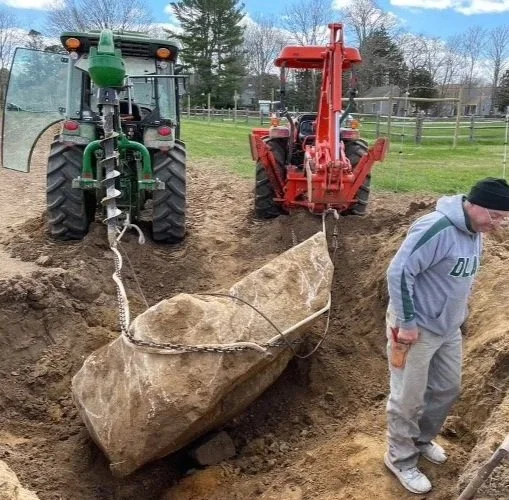 Two tractors are lifting a large boulder from a hole in the ground, with a person standing nearby observing.