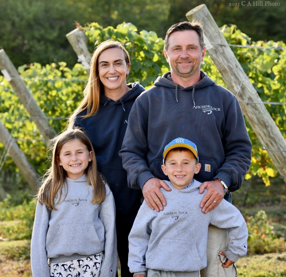 A family of four, two adults and two children, standing outdoors in a vineyard with grapevines in the background. They are all smiling and wearing casual clothing, with the adults and one child wearing matching sweatshirts that say 'Anchor Rock.'