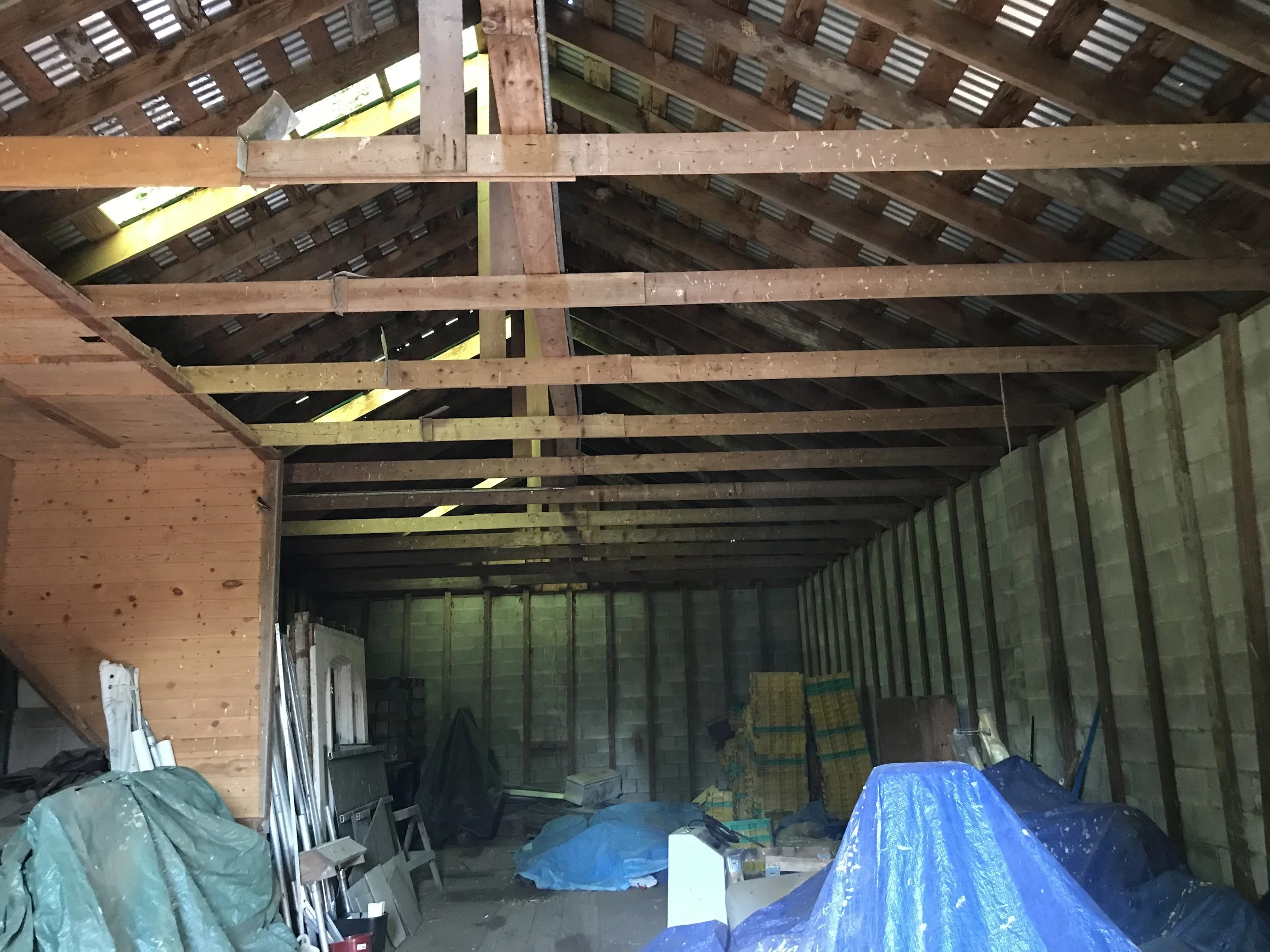 View of the inside of a building under construction with wooden beams, an unfinished roof, and construction materials and covered items on the floor.