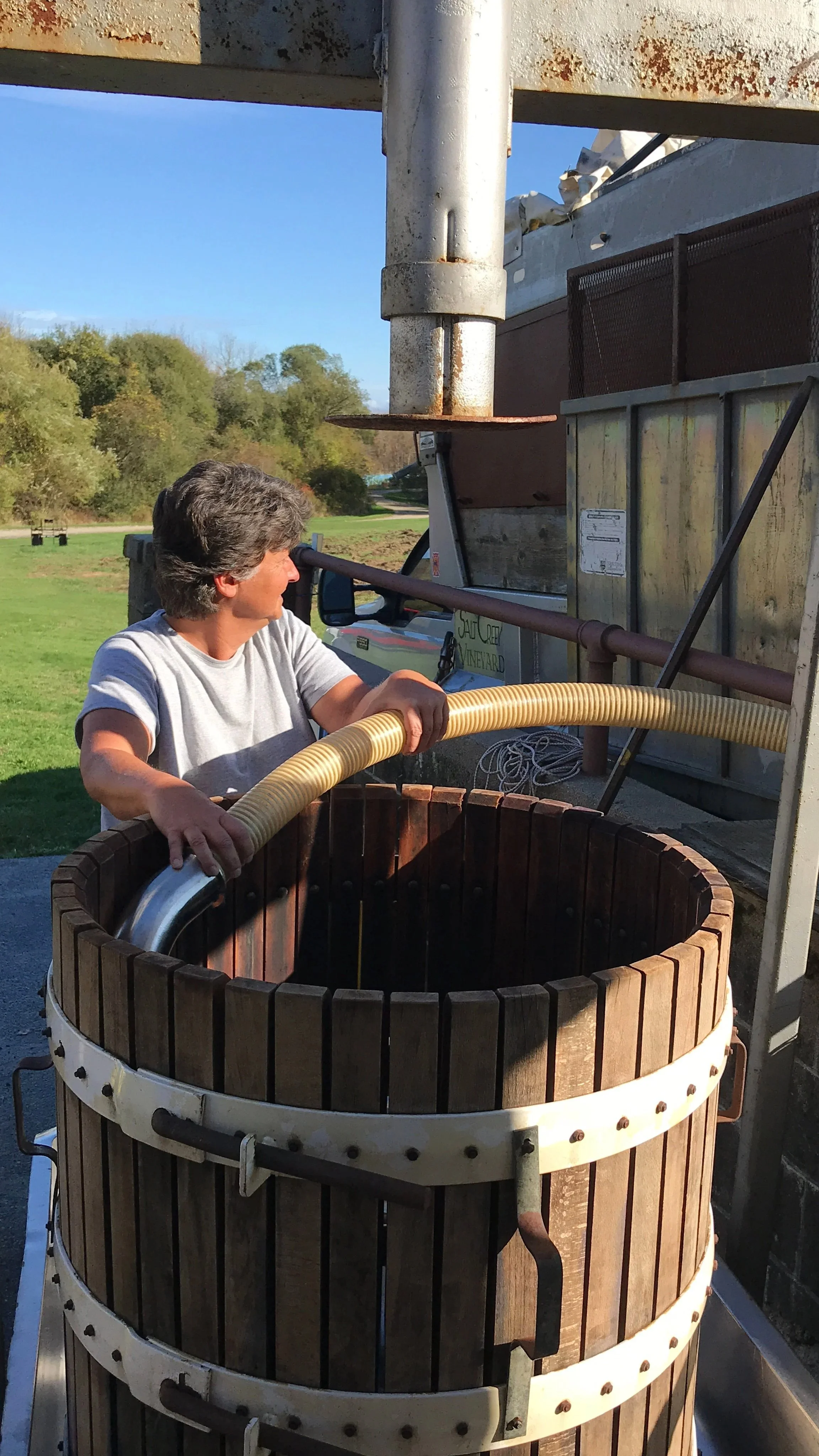A woman operating a grape press outside surrounded by green trees on a sunny day.