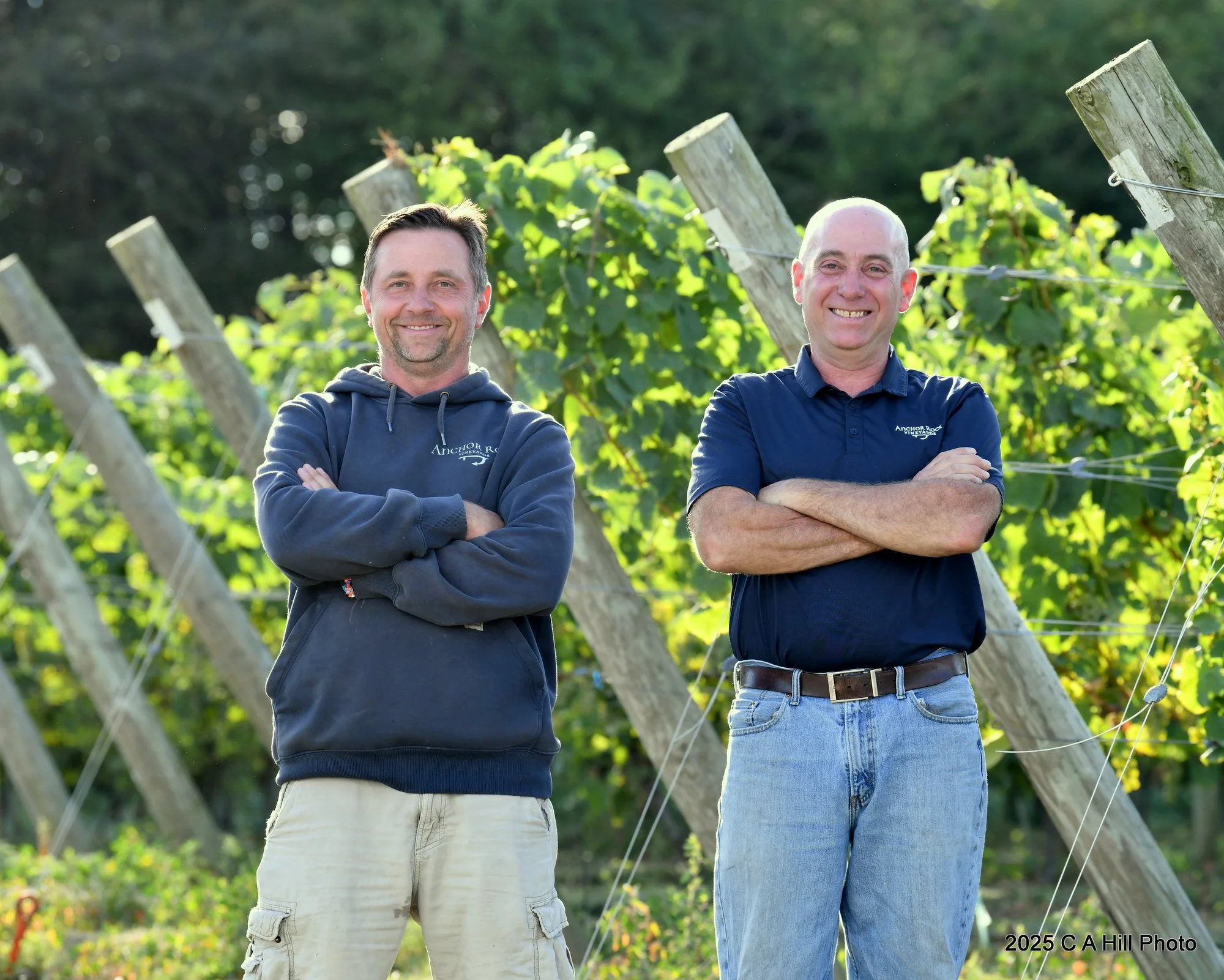 Two smiling men standing outdoors near a vineyard or grapevine trellis, crossing their arms, dressed casually in long sleeves, one in a hoodie and the other in a polo shirt, with trees in the background.