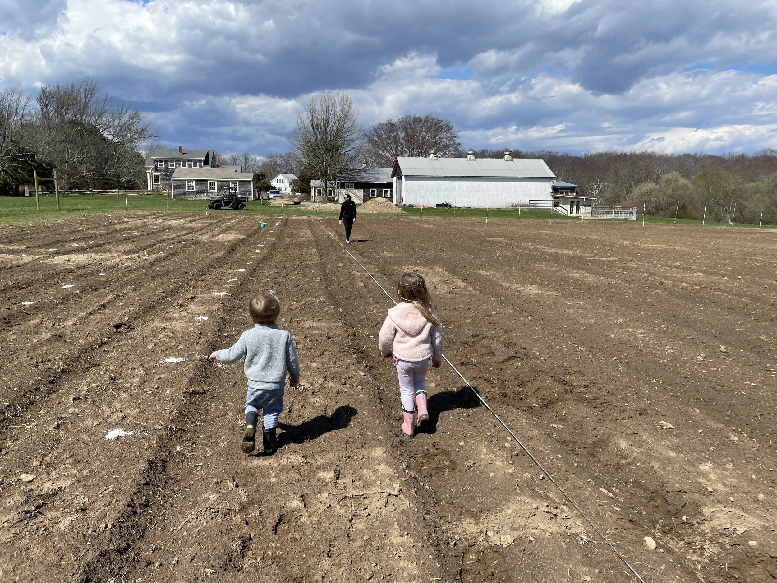 Children and an adult walking across a plowed field on a farm, with farm buildings, trees, and a cloudy sky in the background.