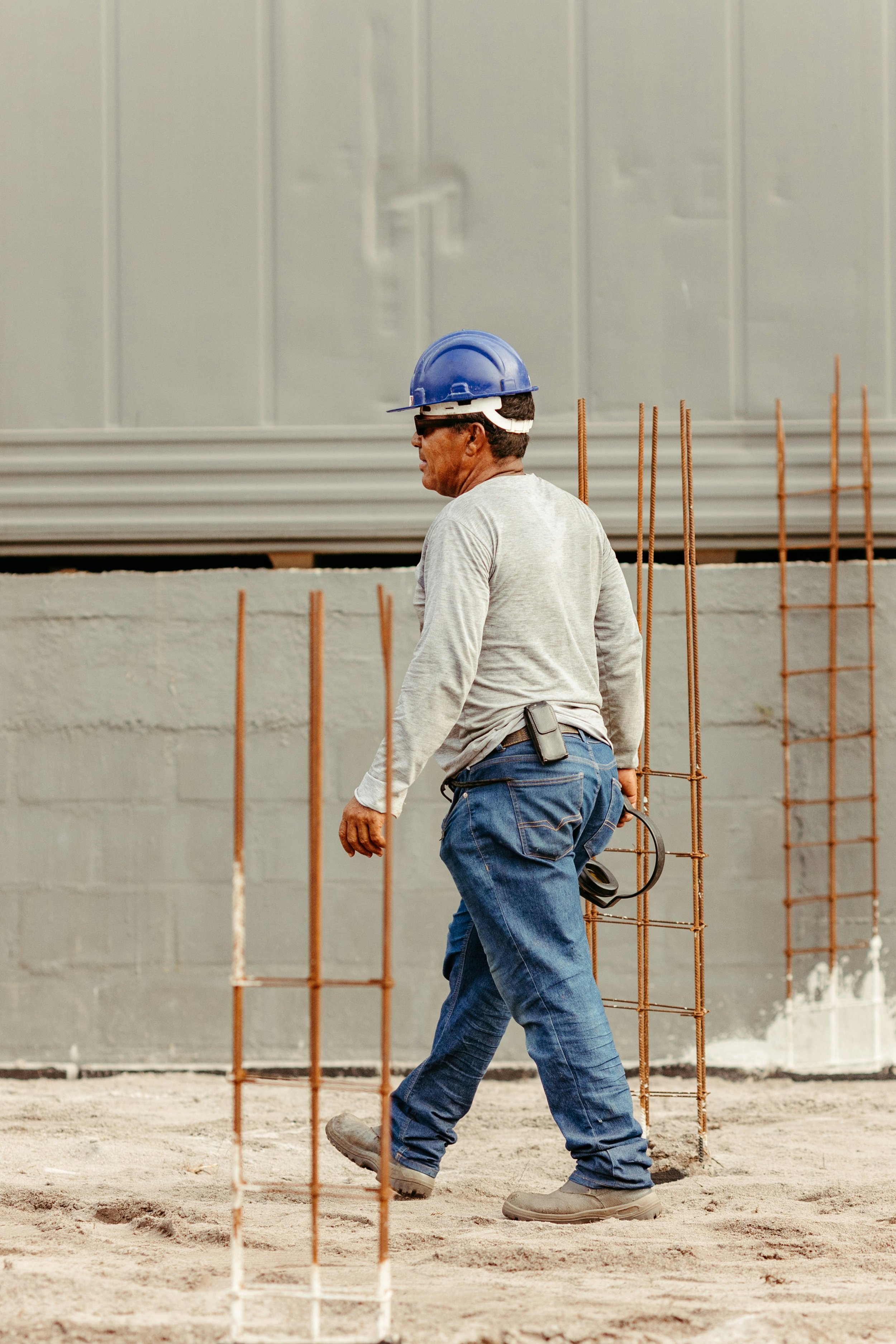 Un trabajador en una obra de construcción usando casco azul, pantalón y botas de trabajo, caminando entre estructuras de metal en un sitio de construcción.