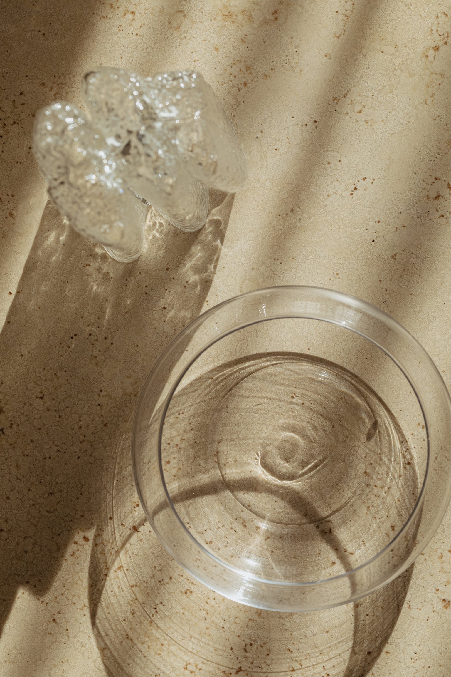 Minimalist glass of water beside artistic glass sculpture casting elegant shadows on a beige textured surface in luxury home setting.