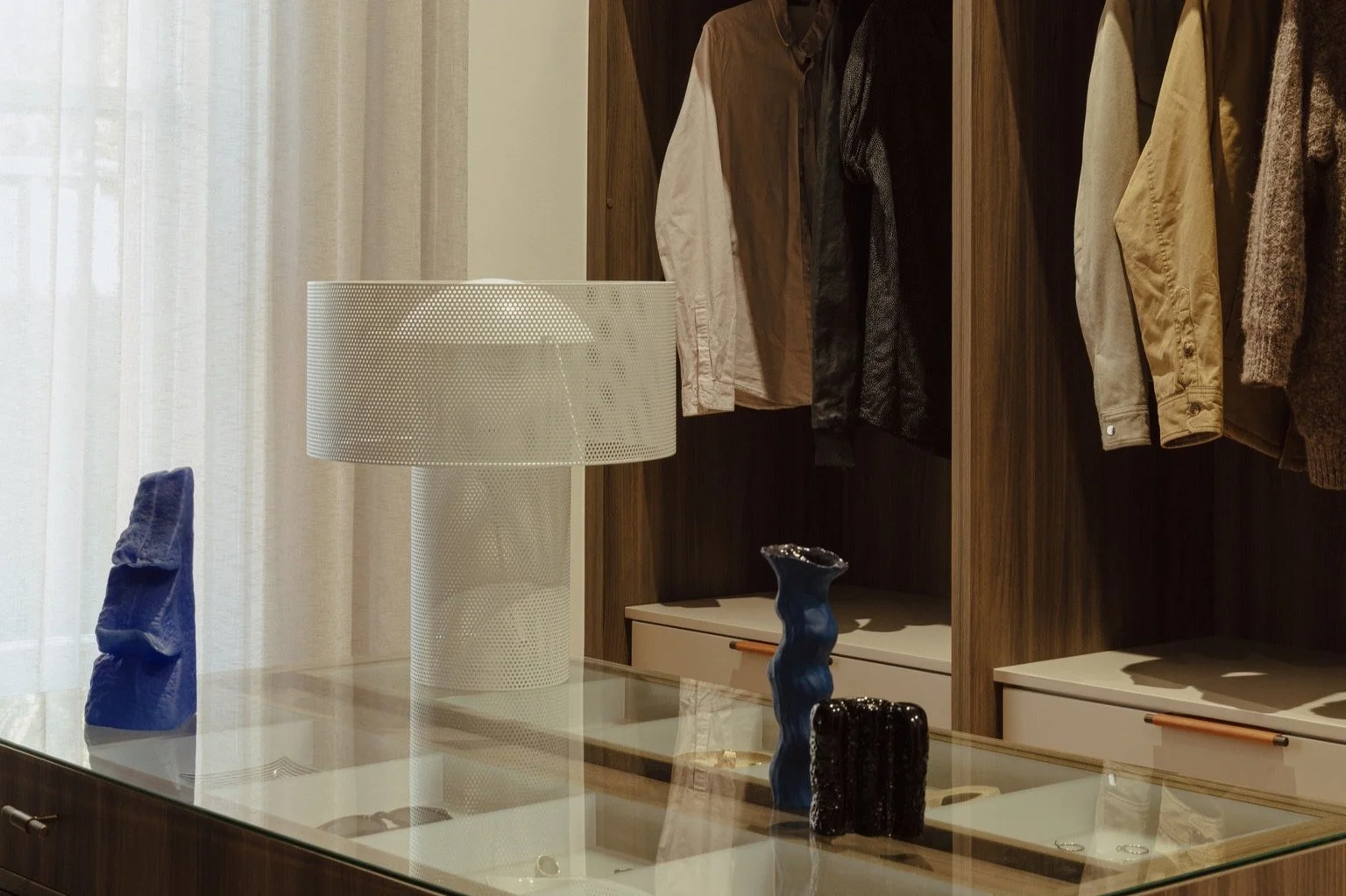 A glass-top display table showcasing three decorative vases in blue, black, and dark blue colors, with a white textured table lamp nearby. Behind the table, there is an open wooden wardrobe containing various hanging jackets and shirts in neutral and dark tones.