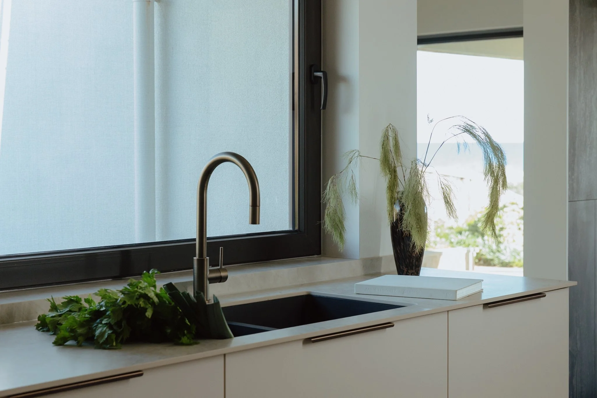 Modern kitchen with beige cabinets, a black sink, a brass faucet, a potted plant with green leaves, a white book, and a tall vase with dried ornamental grass near a large window with frosted glass and a view outside. Bespoke Kitchen Adelaide luxury